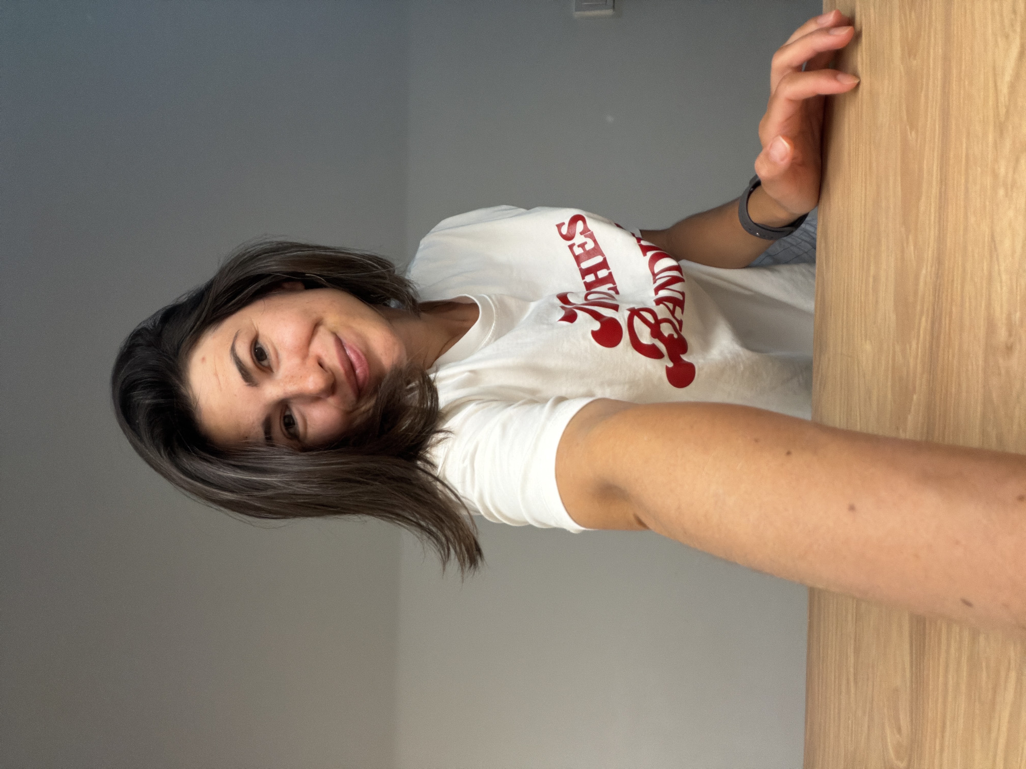 A selfie of a woman with dark hair wearing a white t-shirt with red writing, standing indoors against a grey wall and leaning against a wooden surface. The location is unspecified and there is no visible indication that this photo was taken in Budva, Montenegro.