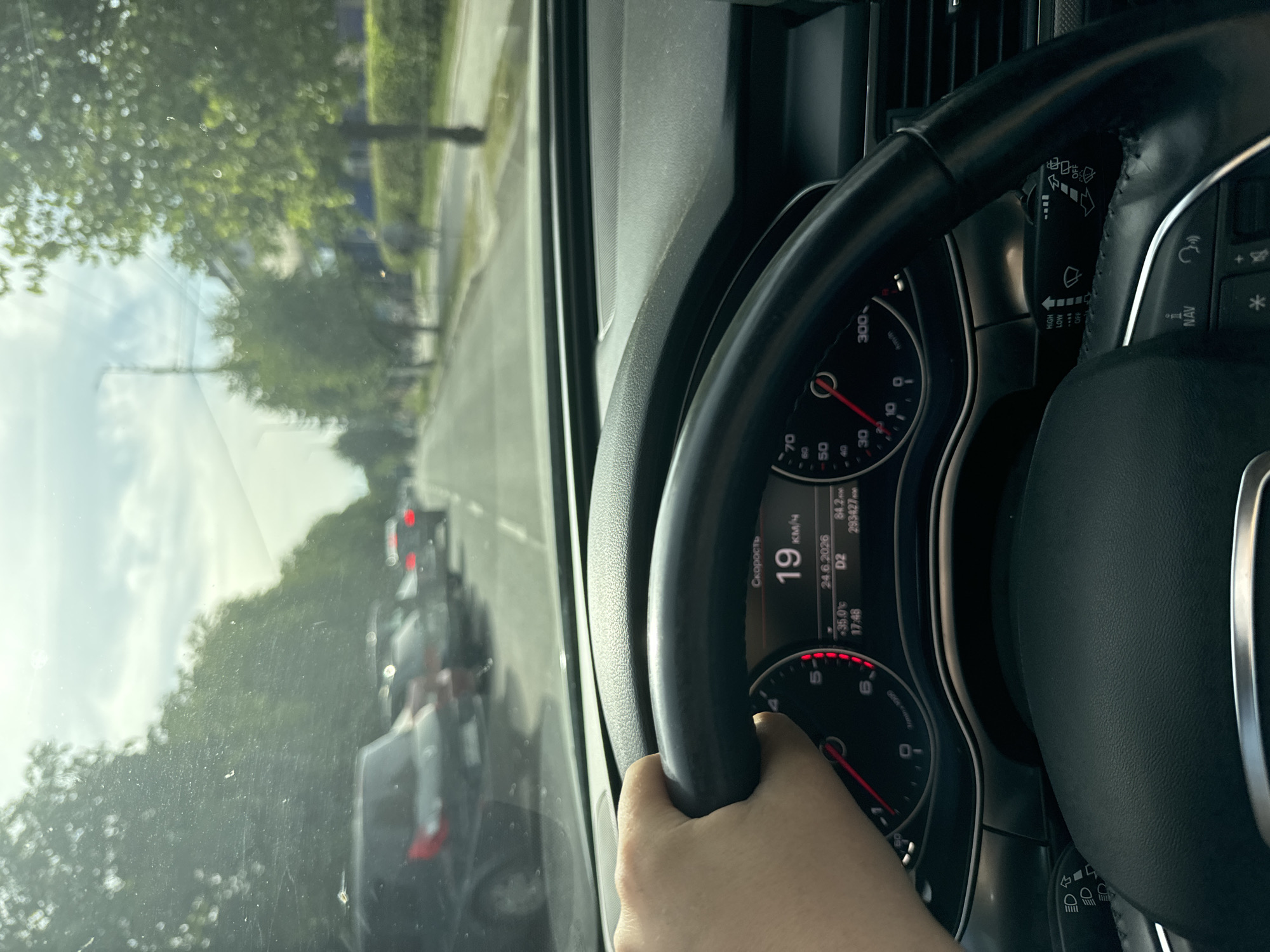 The image shows a person's hand on the steering wheel of a car, driving on a road. The view is from inside the car, looking forward through the windshield. Outside, there are trees and other cars visible, suggesting an urban or suburban setting. The dashboard displays the speed at 19 km/h, the date June 24, 2026, a temperature of 35°C, and the time 17:48. The weather appears to be sunny and warm. The overall scene depicts a typical driving experience. There are also control stalks on the steering column, indicating features like lights and wipers. The image appears to be taken in Sarny, Ukraine, based on the provided location context.
