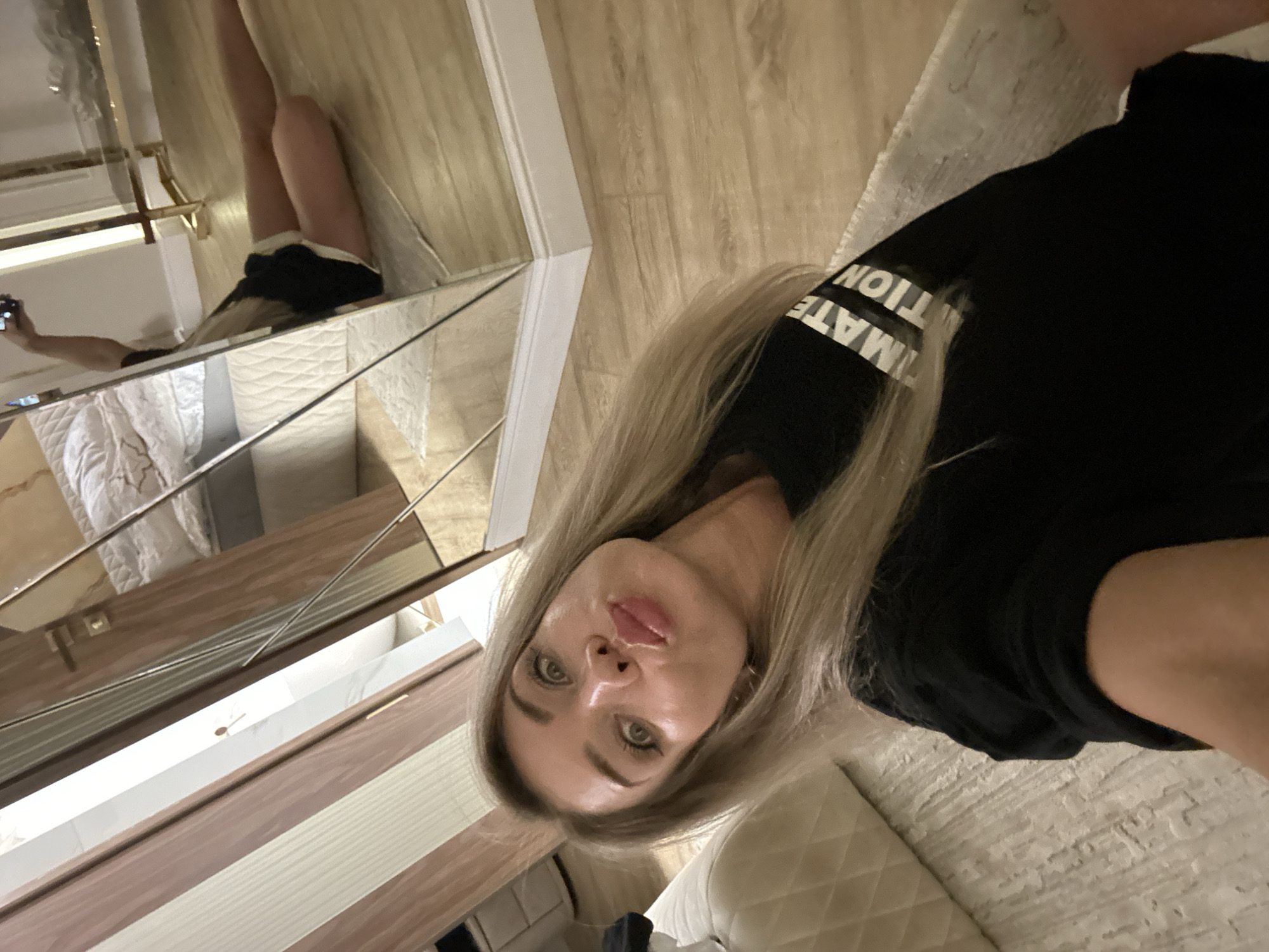 The image is a selfie taken by a woman with blonde hair and heavy makeup. She is wearing a black t-shirt with white text that appears to say "ULTIMATE" and "TION". Her lips are pursed, and she is looking at the camera. She is sitting on a light-colored textured surface, possibly a rug or couch.
In the background, there is a large mirror reflecting another woman who is also taking a selfie with her phone. This reflected woman is wearing black shorts and a black top. The reflection also shows a bedroom with a bed and furniture, and a wooden floor. There are also some mirrored doors in the background.
The image appears to be taken indoors, possibly in a bedroom or dressing area. The lighting is warm and ambient. There are no visible indications of the image being located in Armavir, Russia.