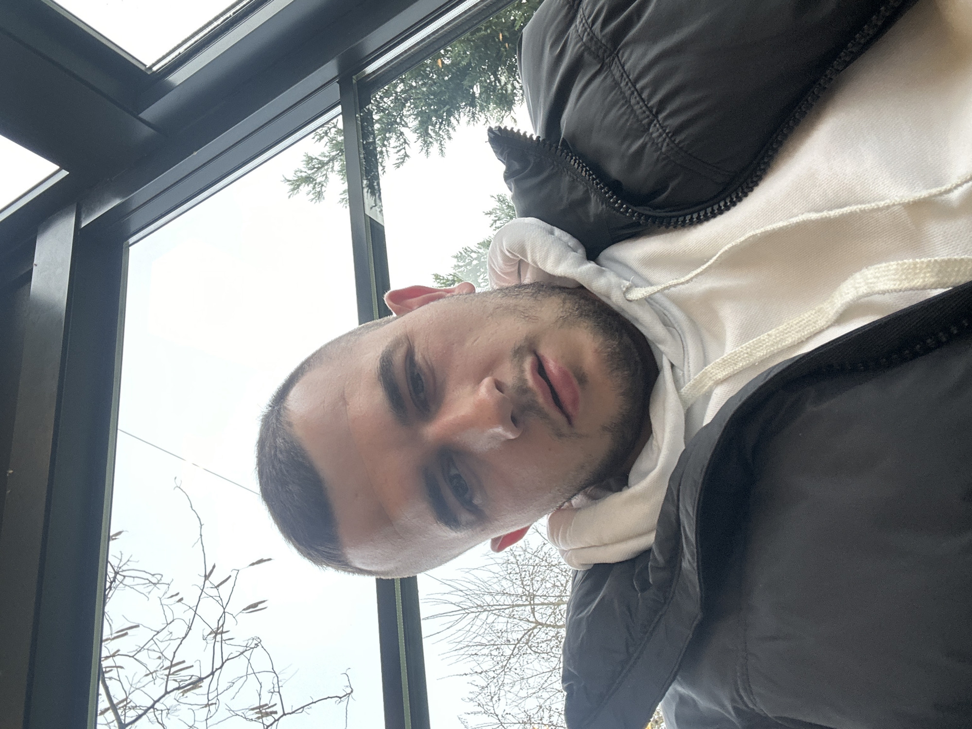 The photograph is a selfie of a Caucasian man wearing a white hoodie and a black puffer jacket. He has short dark hair and stubble. The background suggests he is indoors, possibly near a window, with visible tree branches and a cloudy sky seen through the window. The setting appears to be a building with a glass enclosure.