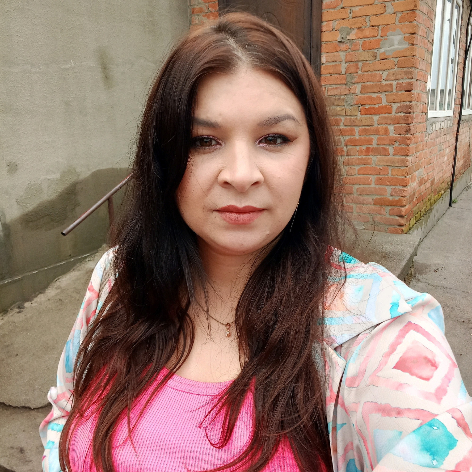 A woman is pictured from the chest up, facing the camera. She has long, dark brown hair that is slightly wavy and falls around her shoulders. She is wearing a bright pink, ribbed tank top and a light-colored jacket with a watercolor-like pattern of pink, blue, and white geometric shapes. A delicate gold necklace with a small pendant is visible around her neck. She has on makeup, including eyeliner and lip color.
The background suggests an outdoor urban or semi-urban setting. To the left, a rough, textured concrete wall is visible, with a metal railing attached. To the right, a brick wall constructed of reddish-brown bricks with visible mortar is present. A portion of a white-framed window with vertical panes is also seen on the brick wall. The ground in the foreground appears to be concrete or asphalt. The overall lighting suggests an overcast day.