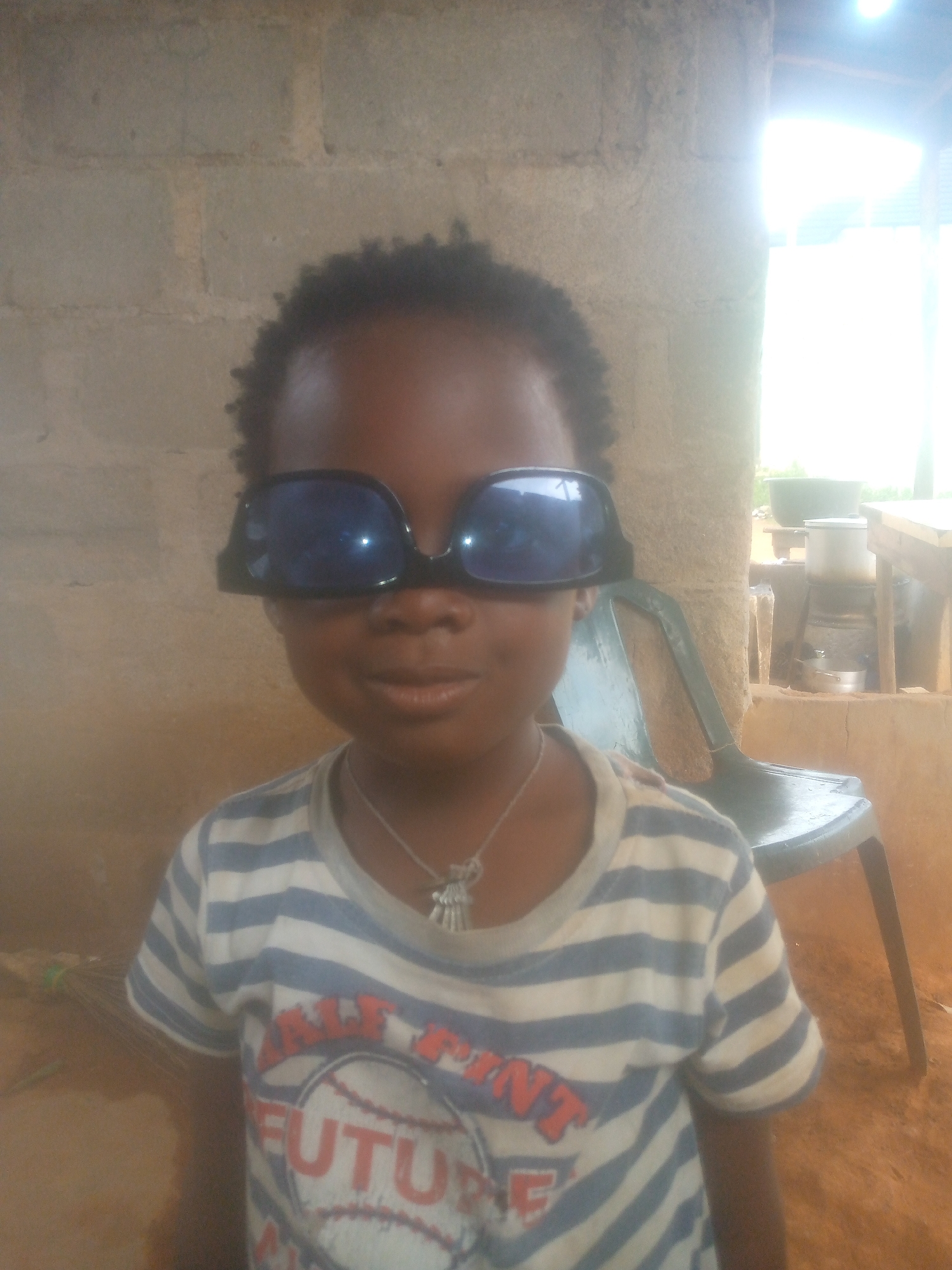A young child with dark skin and short, dark curly hair is the central subject of the image, positioned in a medium close-up, facing directly towards the camera. The child is wearing oversized black sunglasses that cover their eyes and nose, giving them a playful appearance. A slight smile is visible on their face.
They are dressed in a white t-shirt with thin blue horizontal stripes. Around their neck, a silver-colored necklace with small charms hangs. On the front of the t-shirt, a graphic design features what appears to be a baseball. Visible text on the shirt reads "PRINT FUTURE," with the word "FUTURE" being most prominent and in a reddish-orange color.
The background suggests a simple, possibly rural, indoor or semi-outdoor setting. To the left of the child is a light brown brick wall. To the right, a bright, overexposed light source, likely an open doorway or window, illuminates the scene strongly, creating a glare. Behind the child's right shoulder, a dark green plastic chair is visible. Further back, on the right side of the frame, there are some household items including cooking pots and what looks like a makeshift cooking area or table. The ground or floor appears to be an earthy, reddish-brown color. The lighting suggests it is daytime.