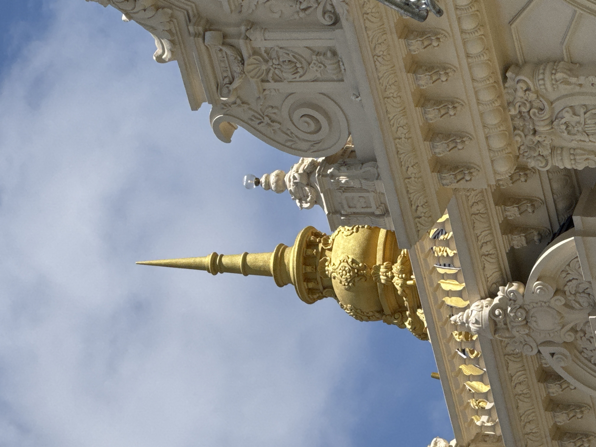 The image shows the exterior architectural details of a building, likely a temple or religious structure, against a blue sky with wispy clouds. Dominating the left side of the frame is a tall, golden spire with intricate decorative elements. To the right of the spire, another smaller spire is visible, topped with a glass orb. The building itself is ornate, constructed from light-colored stone with detailed carvings, including scrollwork, floral motifs, and decorative moldings. A balustrade with golden leaf-shaped elements runs along the top of the visible structure. The overall impression is one of grandeur and intricate craftsmanship. This is a view of the exterior of the Linh Phuoc Pagoda in Da Lat, Vietnam.