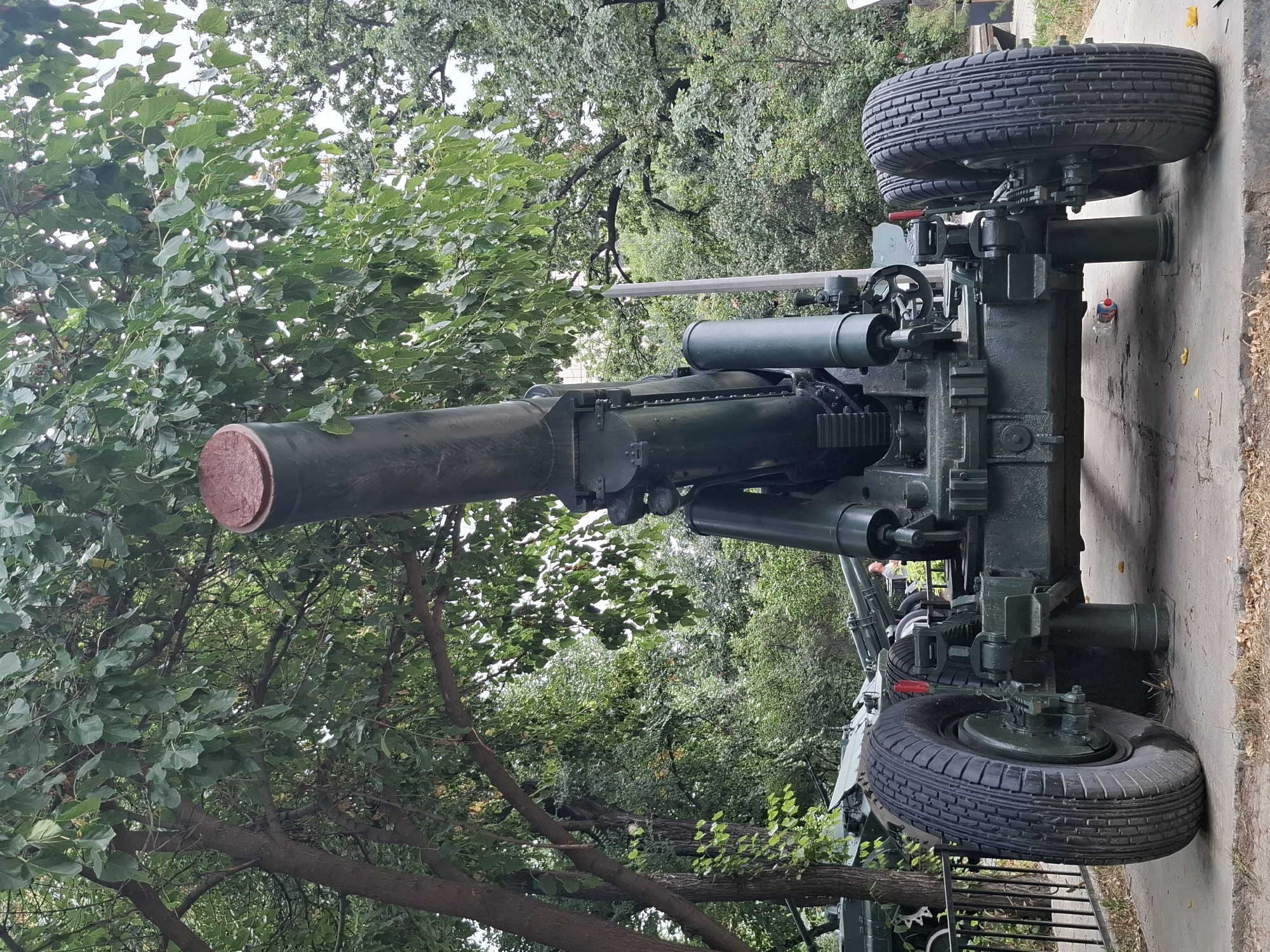 The image shows a large, dark green artillery piece, likely a howitzer or mortar, positioned on a concrete pad outdoors. The weapon is angled upwards and to the left. It has two large, treaded tires on either side of its wheeled carriage. Behind the artillery piece, there is a dark metal fence and further back, numerous trees with green foliage. The sky is overcast. The artillery piece is the primary subject, and its mechanics and large barrel are clearly visible. There are no people clearly depicted in the main view, although a blurred figure can be seen in the background to the right. The overall environment appears to be a park or outdoor exhibition area. The location of the artillery piece in Almaty, Kazakhstan is not discernible from the image itself.