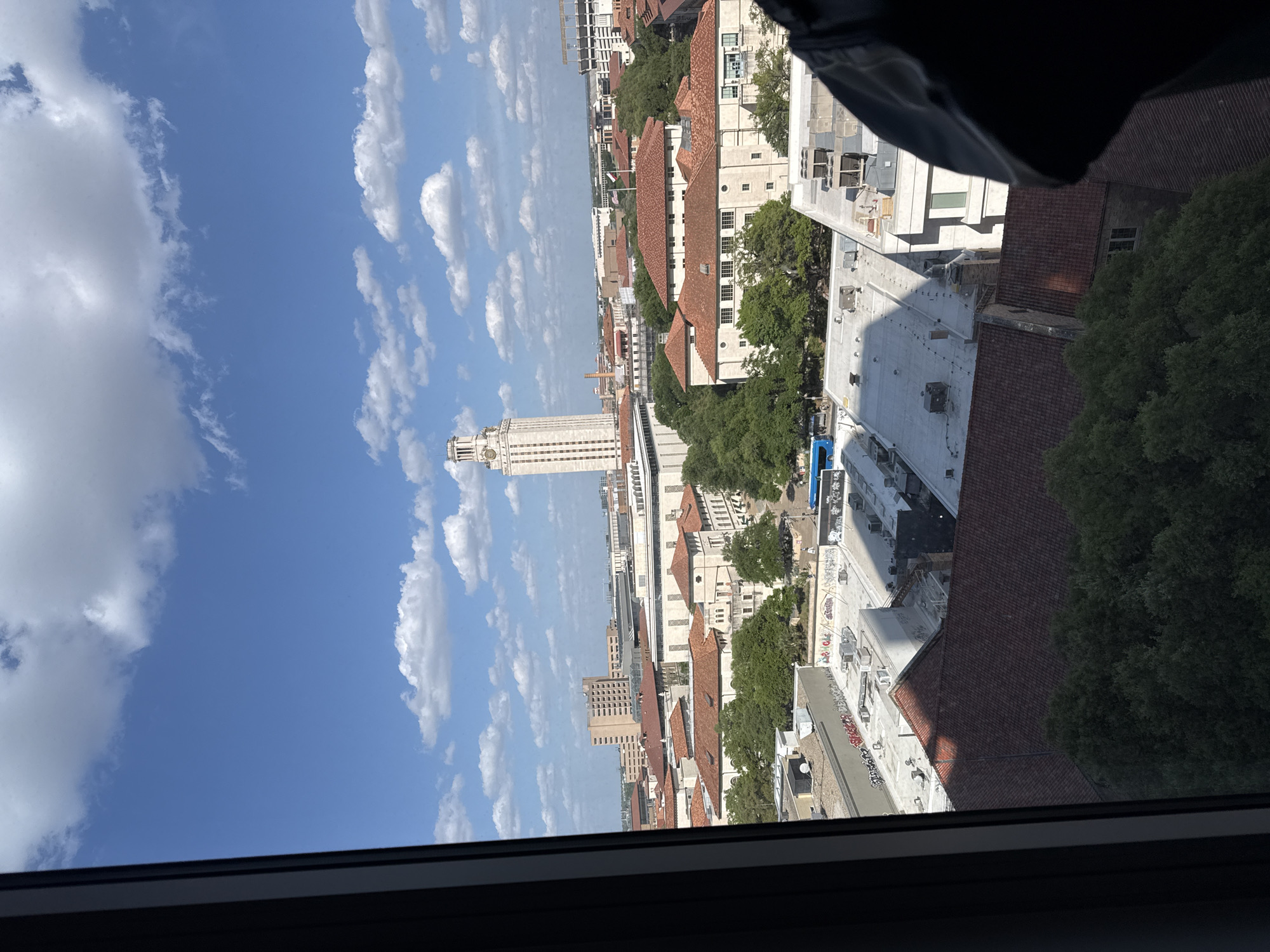 The image depicts a view from an elevated position, likely an office or apartment building, overlooking the city of Austin, Texas. The most prominent landmark is the University of Texas Tower, a distinctive building with a white facade that stands out against the blue sky. The buildings in the foreground and midground have red-tiled roofs and light-colored facades, indicating a university campus or historic neighborhood. The sky is clear with scattered white clouds. The presence of trees lining the streets and rooftops indicates a blend of urban and natural elements in the cityscape. The lower frame of the picture shows the window frame which indicates a view from indoors. The image is oriented vertically, suggesting it was taken with a smartphone.