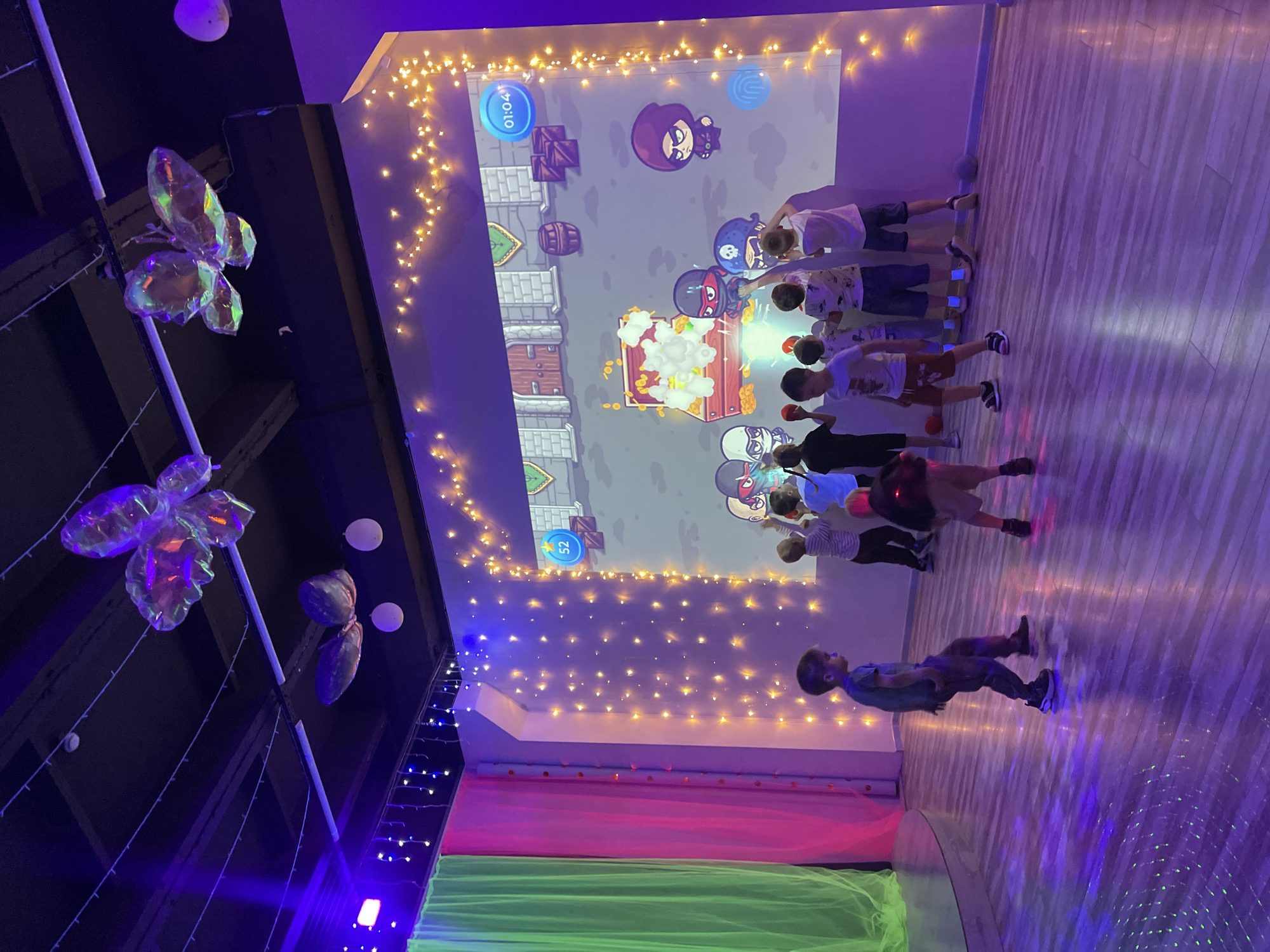 The image depicts an indoor entertainment area, likely a children's party venue or arcade, with a large projection screen displaying a cartoon game. Several children are actively engaged with the projected game, some reaching out towards it. The room is dimly lit, with colored stage lights casting purple and blue hues on the wooden floor, and illuminated by strings of warm white fairy lights adorning the walls and ceiling. Large, translucent butterfly decorations are suspended from the ceiling. On the left side of the frame, draped fabric in green and pink colors adds to the festive ambiance. The children appear to be interacting with a game that features animated ninjas and a treasure chest.