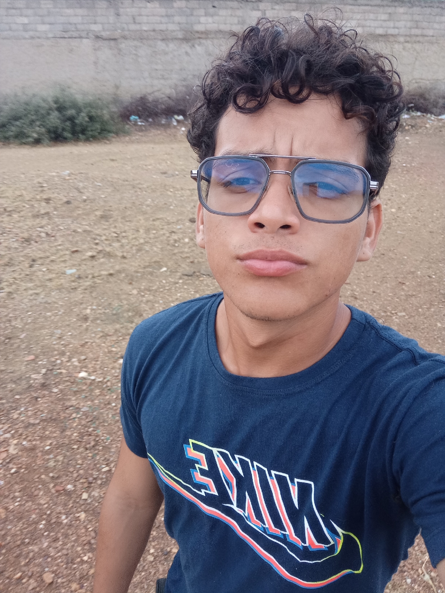 A medium shot portrait shows a young person with curly dark hair wearing a dark blue t-shirt with a stylized colorful "NIKE" logo across the chest. The person is also wearing large, metallic-framed glasses with reflective lenses. The background appears to be an outdoor environment with dry, rocky ground and some sparse vegetation. A low, light-colored brick wall is visible in the upper portion of the image. The scene suggests an informal setting, possibly in a rural or undeveloped area.