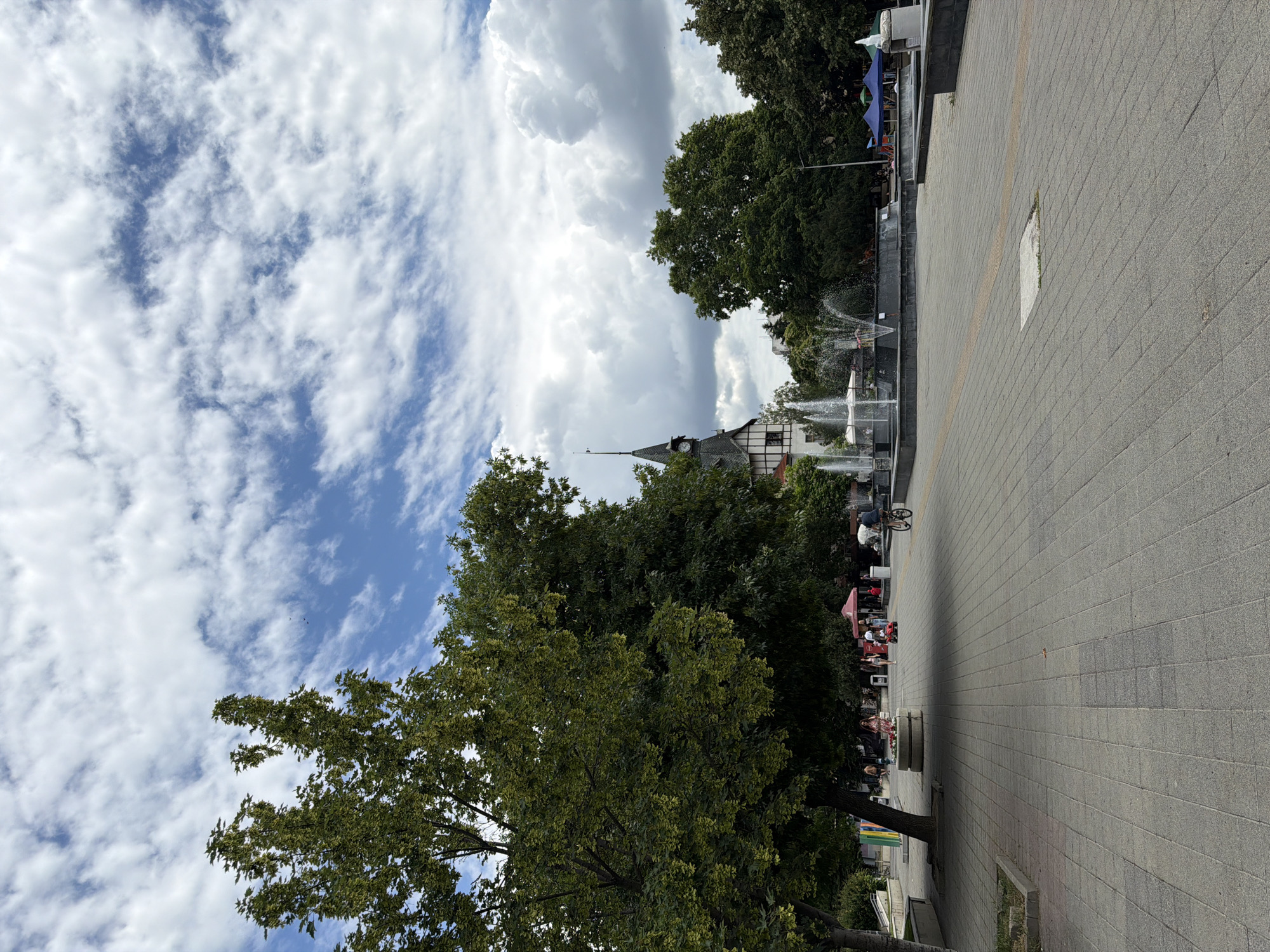 The image depicts a wide, paved pedestrian area in the city of Pazardzhik, Bulgaria, on a cloudy day. In the foreground, the grey paving stones are laid out in a regular pattern, leading towards a fountain in the middle ground. To the left, a large, leafy tree provides shade over a portion of the walkway. Further back, several people are visible, some walking and others gathered near market stalls or benches. A cyclist rides along the path on the right side of the image. In the background, a building with a distinctive dark-colored spire and a clock face is visible, along with more trees and tents. The sky above is filled with a mix of blue sky and white, puffy clouds. The overall scene suggests a public space in a city, likely a park or a central square, with a moderate amount of activity.