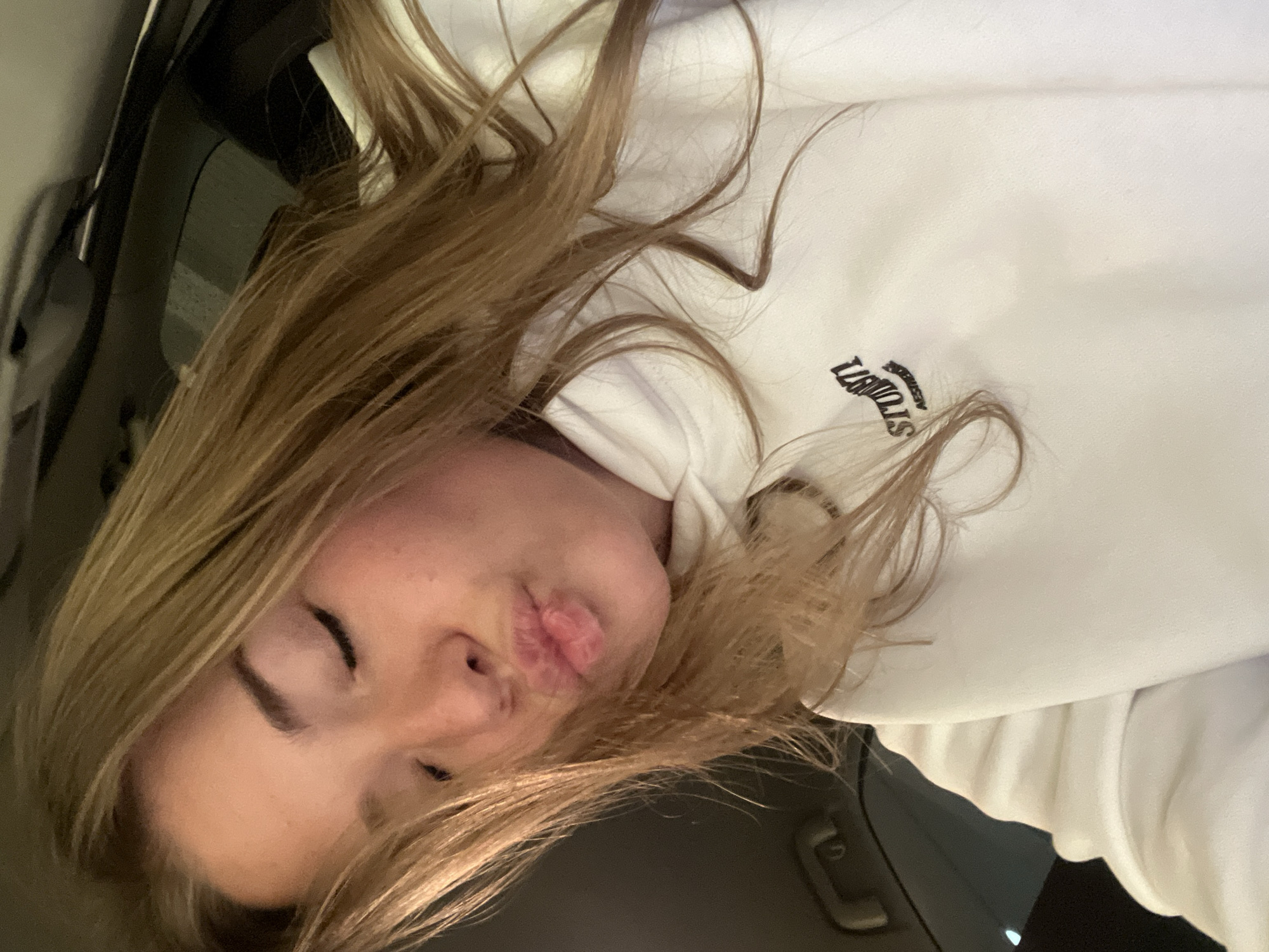 An image depicts a young female inside a vehicle, presumably a car, in Budva, Montenegro. The subject has long, light brown hair styled with a center part and is shown from the chest up, angled slightly. Her eyes are closed, and her lips are pursed forward in a "kiss" gesture. She is wearing a white hooded sweatshirt with a dark-colored logo visible on the left chest area, partially obscured by hair, displaying text that appears to include "STUON71" and "AESTHETIC". The background shows the dark interior of a vehicle, with a light-colored headliner and a dark grab handle visible in the upper right quadrant of the image. The lighting is soft and appears to be internal to the vehicle.