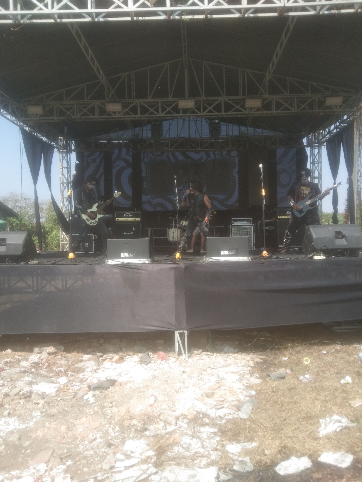 Three male musicians perform on an outdoor stage in Kecamatan Haurgeulis, Indonesia. The musicians are dressed in dark, elaborate costumes and face paint, resembling a rock band. The musician on the left is playing a bass guitar. The musician in the center is singing into a microphone and wearing a vest over a dark shirt. The musician on the right is playing a guitar. The stage is set up with amplifiers, speakers, and a large screen displaying a graphic pattern. Black drapery hangs from the sides of the stage. The ground in front of the stage is littered with debris, including rocks, broken concrete, and trash. The overall scene suggests an outdoor music event.