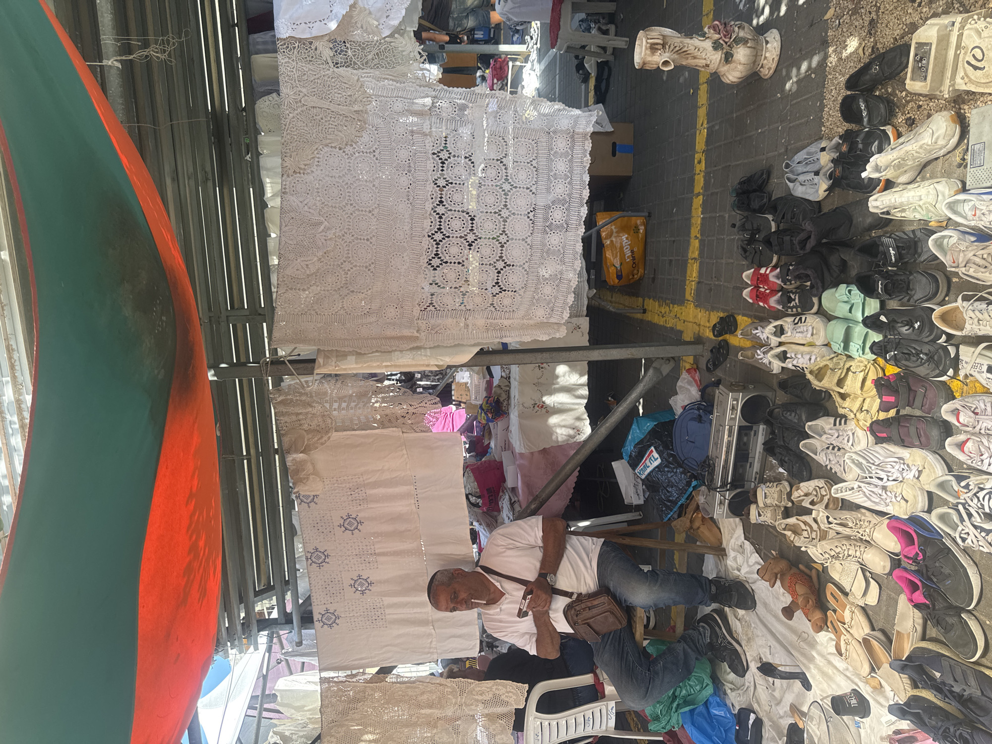 A man sits on a chair at a market stall displaying various items, including crochet items, shoes, and other goods. He wears a white t-shirt, jeans, and a leather shoulder bag. A large orange and green awning is partially visible in the top left corner of the frame. The flooring suggests an outdoor market setting. There are items such as a boombox, handbags, and other household goods scattered on the ground around the shoes. Based on the products being sold in the stalls, the scene takes place in a flea market, likely in Tel-Aviv, Israel.