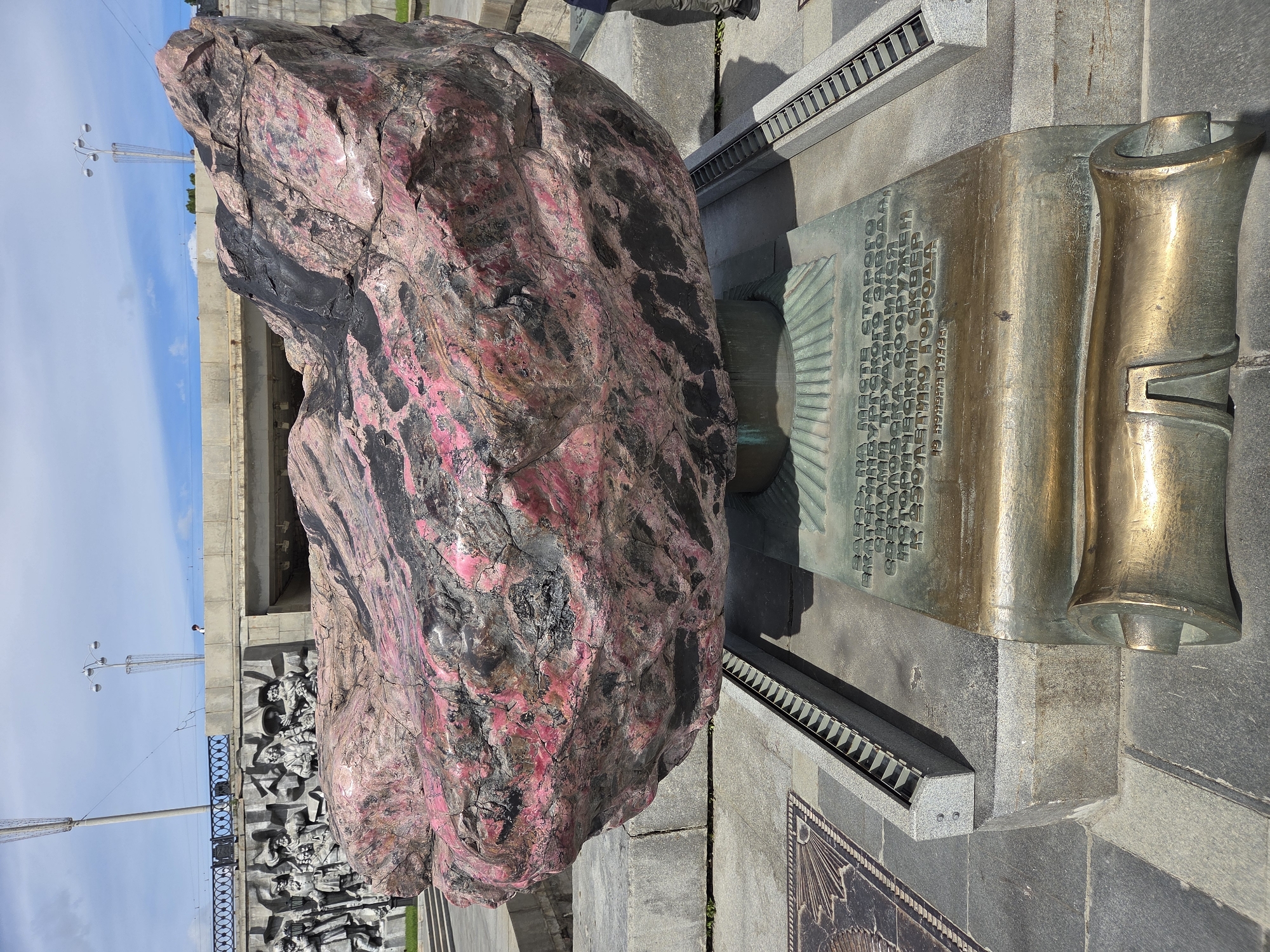 This is a ground-level, eye-level, medium shot of a large, rough pink and black boulder monument in Yekaterinburg, Russia, situated on a granite base with a bronze plaque. The boulder is positioned centrally, appearing to float above the plaque. The plaque is shaped like a scroll, with text in Cyrillic script. Behind the boulder, a relief sculpture depicting historical figures is visible on a grey wall, with a bridge and streetlights in the background. The sky is partly cloudy, and the ground is paved with grey stone tiles. A metal grating runs along the edge of the monument's base. There are no people prominently featured in the image.