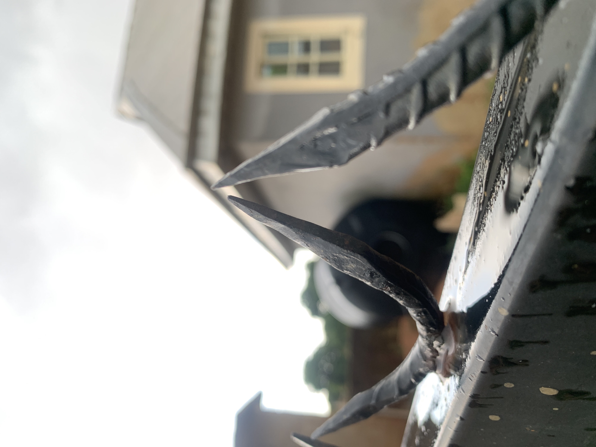 The image shows a close-up of a black metal fence or gate with sharp, pointed spikes on top. The metal is wet, possibly from rain. In the background, a blurred building with a window is visible. Based on the city provided, the image is most likely located in Farin Yashe, Nigeria. There are no people visible in the image. The focus is on the fence as a security measure. The environment appears to be residential.
