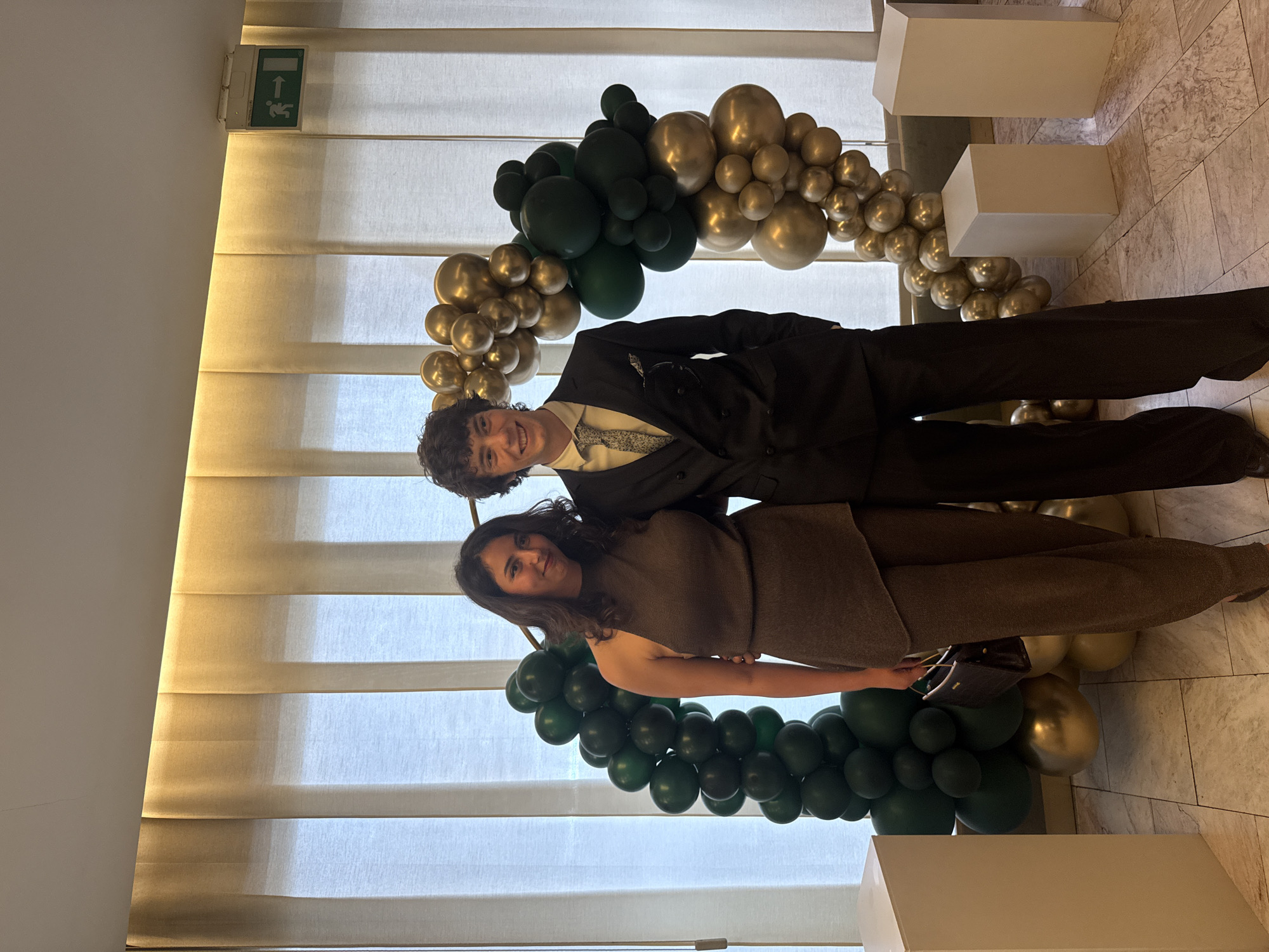 Two people stand in front of a semi-circular balloon arch. The balloon arch consists of gold and green balloons. The man is wearing a suit and the woman is wearing a dress. There are three white rectangular boxes visible in the image. The background consists of light colored blinds and an exit sign. The location is in Sofia, Bulgaria.