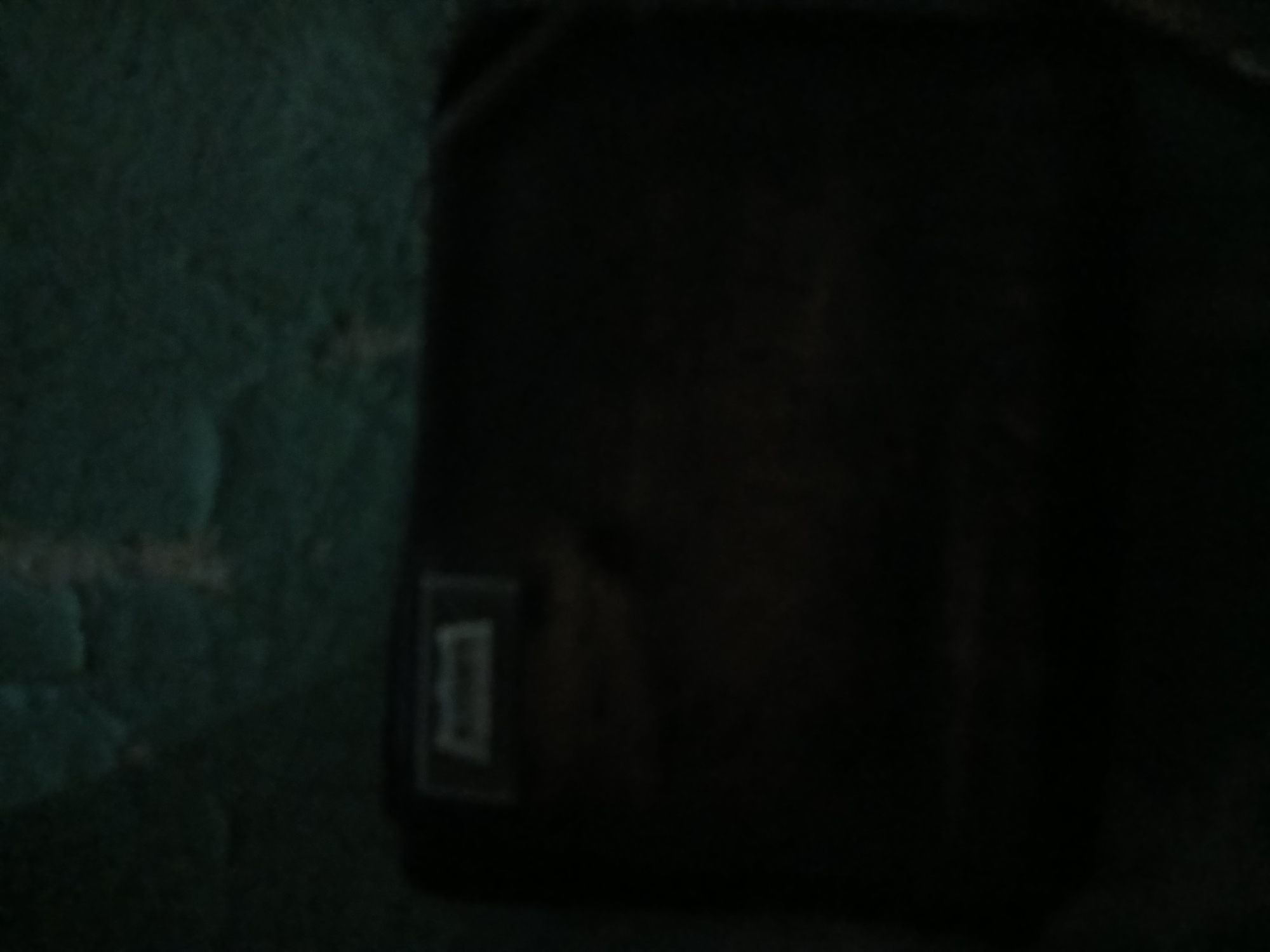 The image depicts a dark, grainy scene, likely captured in low light conditions. The primary subject is a dark, rectangular object, possibly a book or a box, situated on the left side of the frame. A faint, lighter rectangular emblem is visible on the surface of this object. The background is a dark, textured surface, appearing green or teal, with some lighter, irregular patches suggesting a rough or porous material. The overall atmosphere is dim and somewhat indistinct. There is no information within the image to suggest a specific location in Kademangan Kulon, Indonesia, or any identifiable people, actions, or interactions.