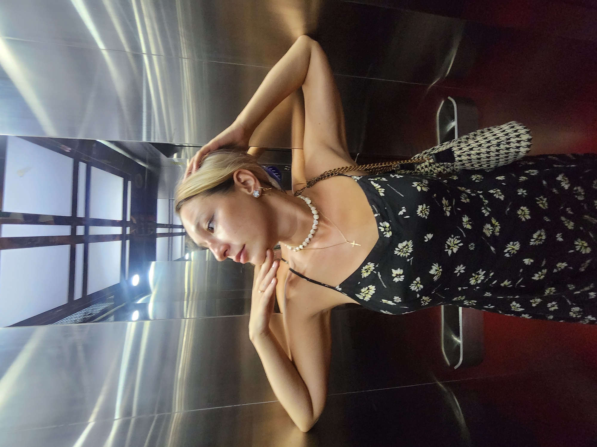 A young woman with blonde hair is standing in an elevator. She is wearing a black dress with a floral pattern and a pearl necklace with a cross pendant. Her right hand is on her head, and her left arm is bent, with her hand resting on her chin. A black and white patterned handbag with a gold chain is slung over her right shoulder. The elevator walls are made of brushed metal, and the ceiling has a grid of fluorescent lights. The floor of the elevator is a dark red. The woman is looking to her left, with a slight smile. The scene is static, with no apparent action beyond the woman's pose.