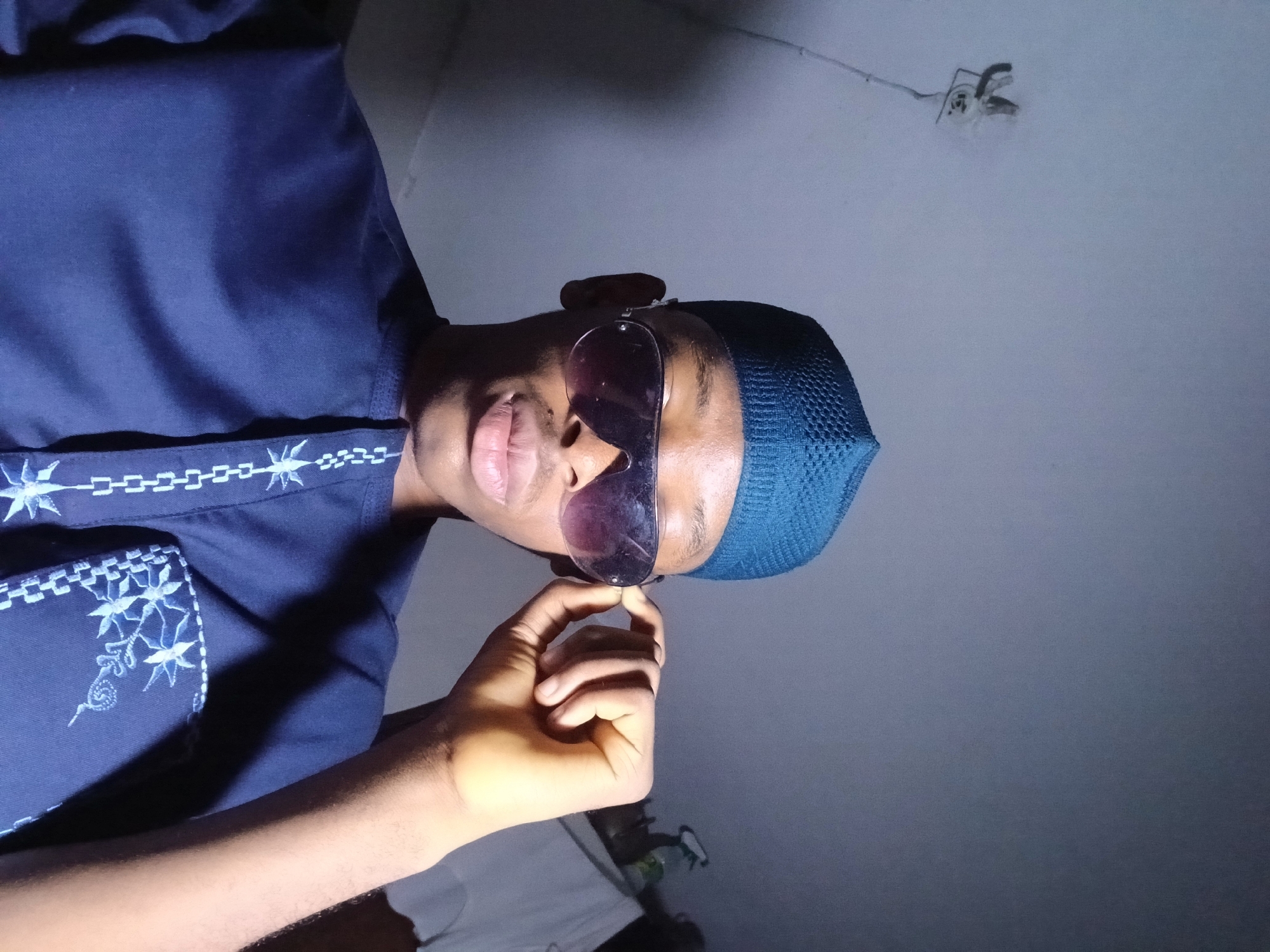 A medium, low-angle shot depicts a person of color wearing a dark blue kufi cap and sunglasses. The person is also wearing a dark blue shirt with embroidered details around the collar and pocket. The person is holding their sunglasses frame between their thumb and index finger. The background is a dimly lit room with a white ceiling. A light fixture can be seen on the ceiling. The photograph was taken in a dark area; it is impossible to determine if the person is within the city of Lagos, Nigeria.
