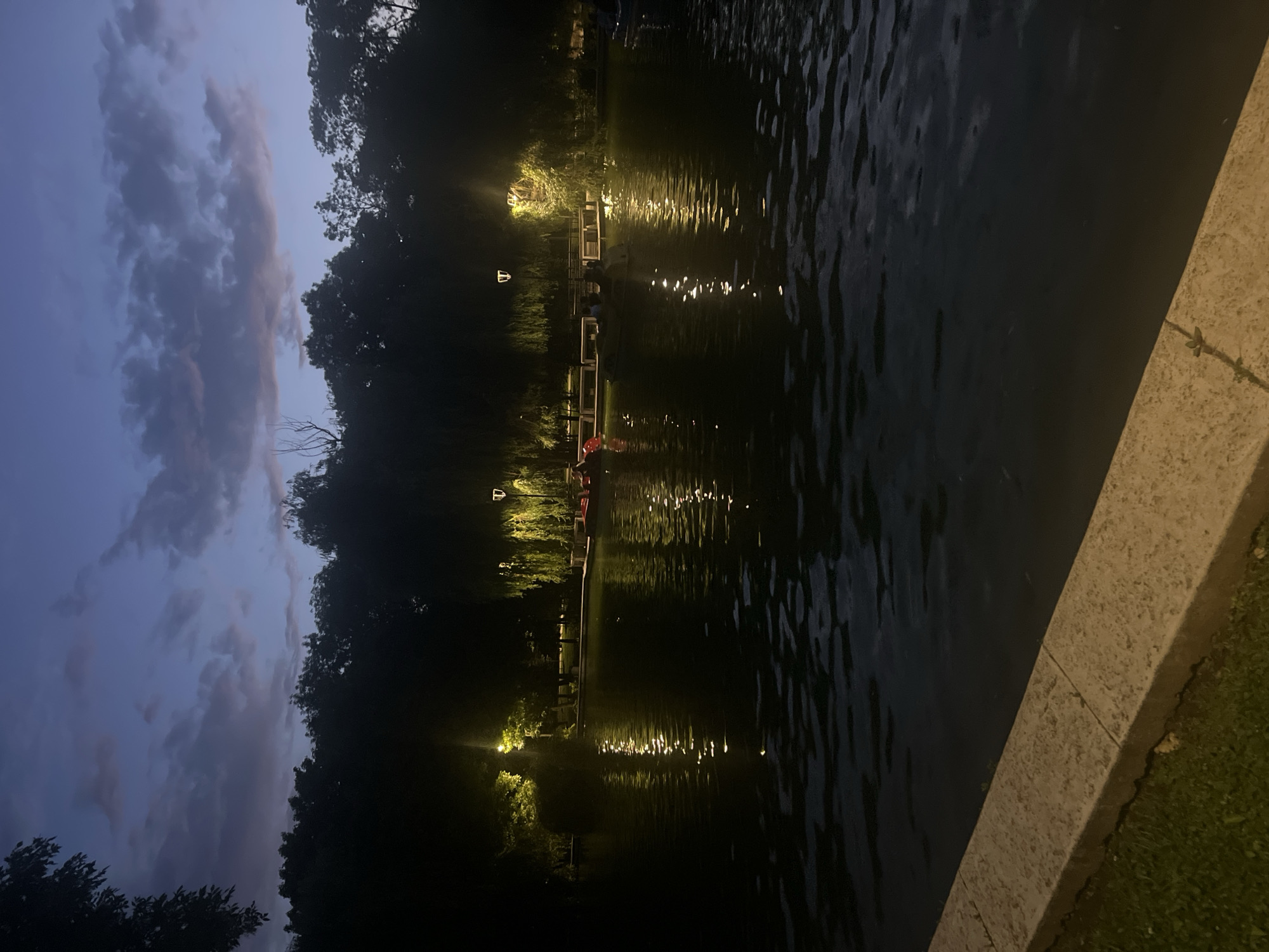 This image depicts a serene evening scene by a body of water, likely a lake or a large pond, at dusk or early night in Dobrich, Bulgaria. The sky at the top is a deep twilight blue, streaked with elongated, soft grey clouds.
In the foreground, a textured, light-colored concrete or stone edge runs diagonally, separating the dark, rippling water on the left from a narrow strip of green grass on the right, which is partially illuminated by a warm light source just outside the frame on the right.
Across the water, the far bank is lined with dense, dark silhouettes of trees. Multiple artificial lights are visible along this bank, some nestled within the foliage, casting a warm yellow-orange glow on the trees and creating prominent, shimmering reflections across the water's surface. A wooden or light-colored pier or walkway extends along the far bank, illuminated by these lights. Several indistinct figures can be seen on the pier, and what appear to be red small boats or floats with figures are visible in the water near it. An isolated, square-shaped white light fixture hangs above the water in the mid-distance.
The overall atmosphere is calm and tranquil, with the artificial lights providing a striking contrast to the dim natural light of the evening. There is no visible text in the image.