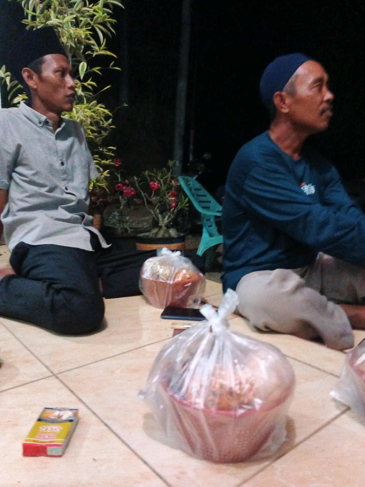 Two men are seated on a tiled floor outdoors at night, in what appears to be a residential setting in Brangan Mulya, Indonesia. The man on the left, wearing a black peci and a light-colored collared shirt with dark pants, is looking to his right. The man on the right, wearing a dark blue peci and a blue long-sleeved shirt with light-colored pants, is looking to his right. In front of the man on the left is a potted plant with green foliage and red flowers. In front of the men are two plastic-wrapped bowls, presumably containing food. A cigarette pack is lying on the tiles to the left of the bowls. A green plastic chair is visible in the background behind the man on the right. The overall lighting is dim, suggesting it is nighttime.