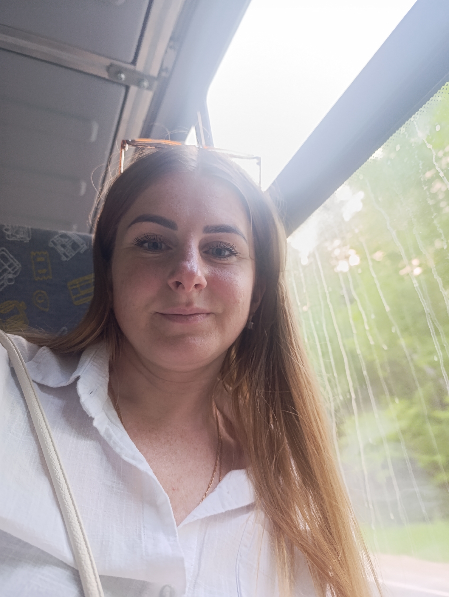 A medium shot captures a woman inside a vehicle, likely a bus or train, given the window and seat visible in the background. The woman, positioned on the left side of the frame, has fair skin, light brown hair, and blue eyes. She wears a white button-down shirt and a thin gold necklace. Sunglasses rest on her head.
The background shows a bus seat with a yellow and grey pattern, and a window with condensation or water droplets obscuring the view outside. Light enters through the window, creating a bright area. The location appears to be in the city of Unknown, Unknown, judging by the details visible through the bus window.