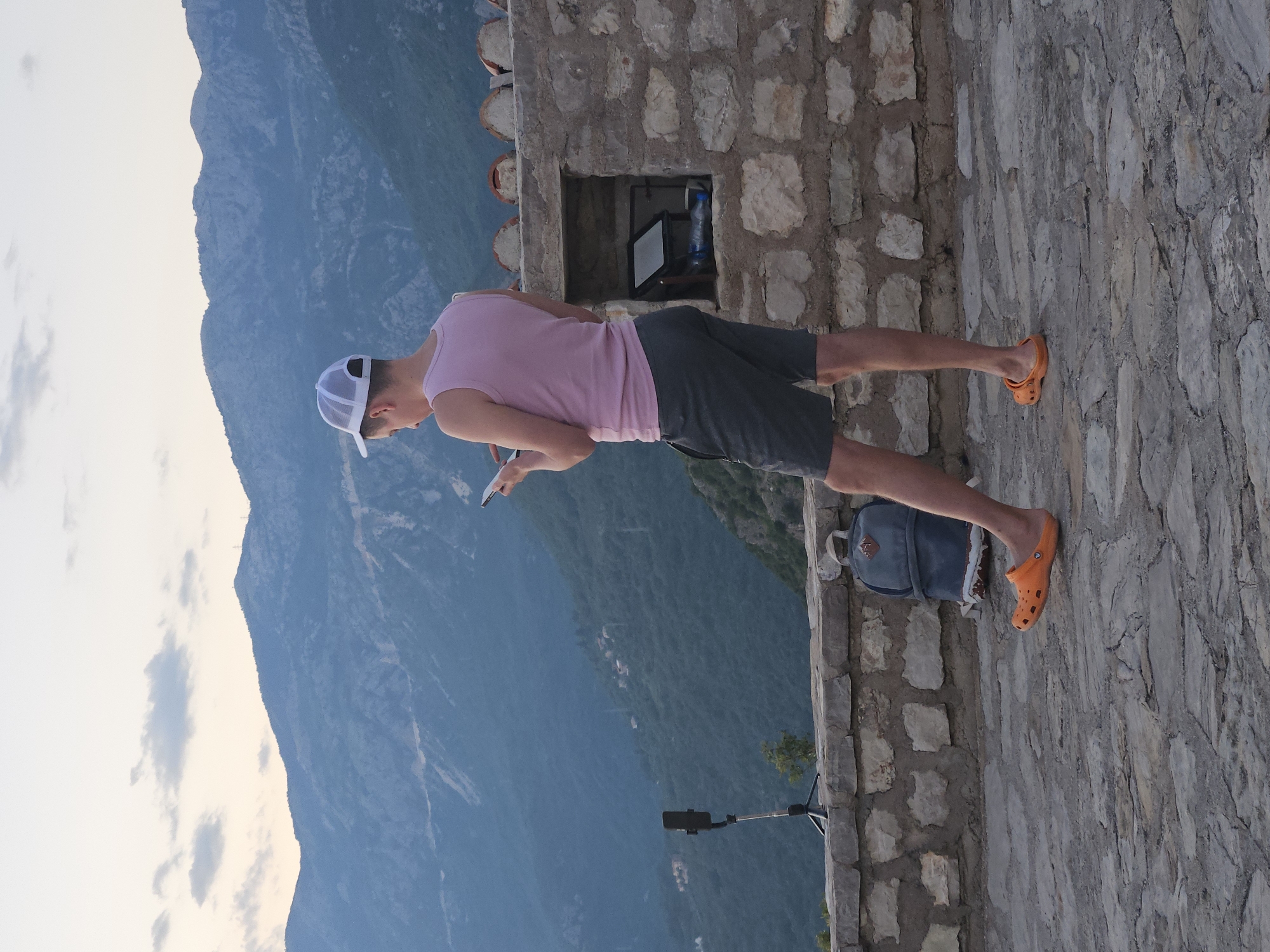A person wearing a pink tank top, dark shorts, a white trucker hat, and orange crocs stands on a stone-paved area, holding a smartphone. The person is looking down at the phone. Behind the person, a stone wall with a small alcove containing a tablet and a water bottle is visible. A blue backpack rests against the wall. To the left, a tripod with a phone attached stands on the wall. The background features a vista of layered, hazy blue mountains under a pale sky. The scene appears to be an overlook in Sveti Stefan, Montenegro, given the architectural style of the wall and the mountainous coastal landscape.