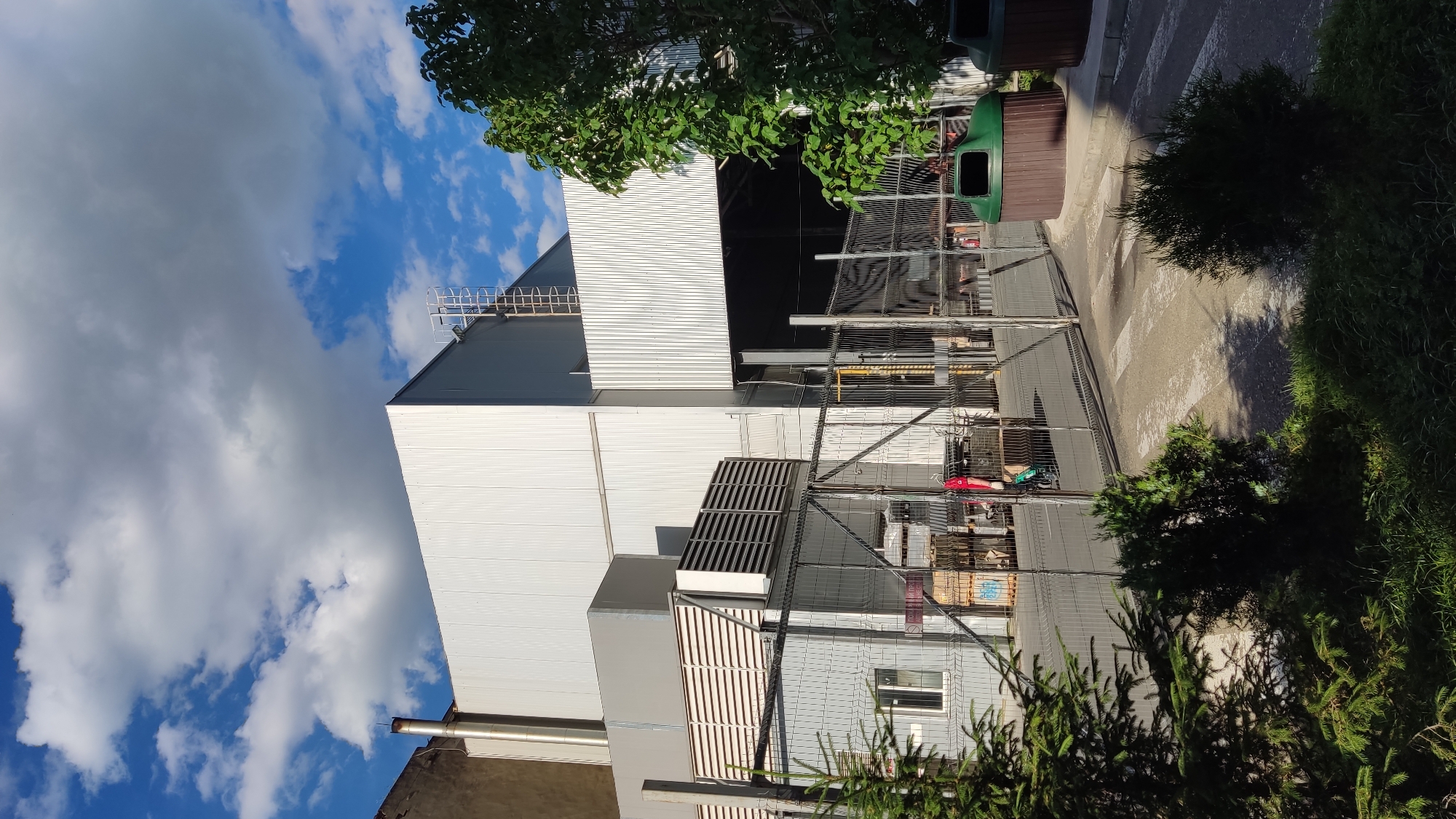 The image depicts an outdoor scene of an industrial or commercial area in Krasnodar, Russia, under a bright, partially cloudy sky.
The main subject is a large, multi-story building, likely a factory or warehouse, featuring light-colored corrugated metal siding. A ladder for rooftop access is visible on one section of this structure. On a lower part of the building, two rectangular ventilation grates or air conditioning units are mounted. A small window is also visible on a lower building section.
A metal fence or barrier runs across the middle ground, separating the building complex from a paved area. Behind this fence, various items are stored, including what appear to be stacked pallets or crates and some indistinct red objects. In front of the fence, there is a paved pathway or road featuring clear white zebra crossing markings.
To the right and in the foreground of the image, there is an abundance of green foliage, including both coniferous trees and other bushes, casting shadows on the ground. Two green and brown public bins or dumpsters are situated near the paved area and the fence. The sky above is a mix of bright blue and white fluffy clouds, with some darker grey clouds also present.
No people are visible in the scene. The lighting indicates it is daytime, likely mid-morning or afternoon, given the clear sunlight and shadows. There is no discernible text visible in the image.