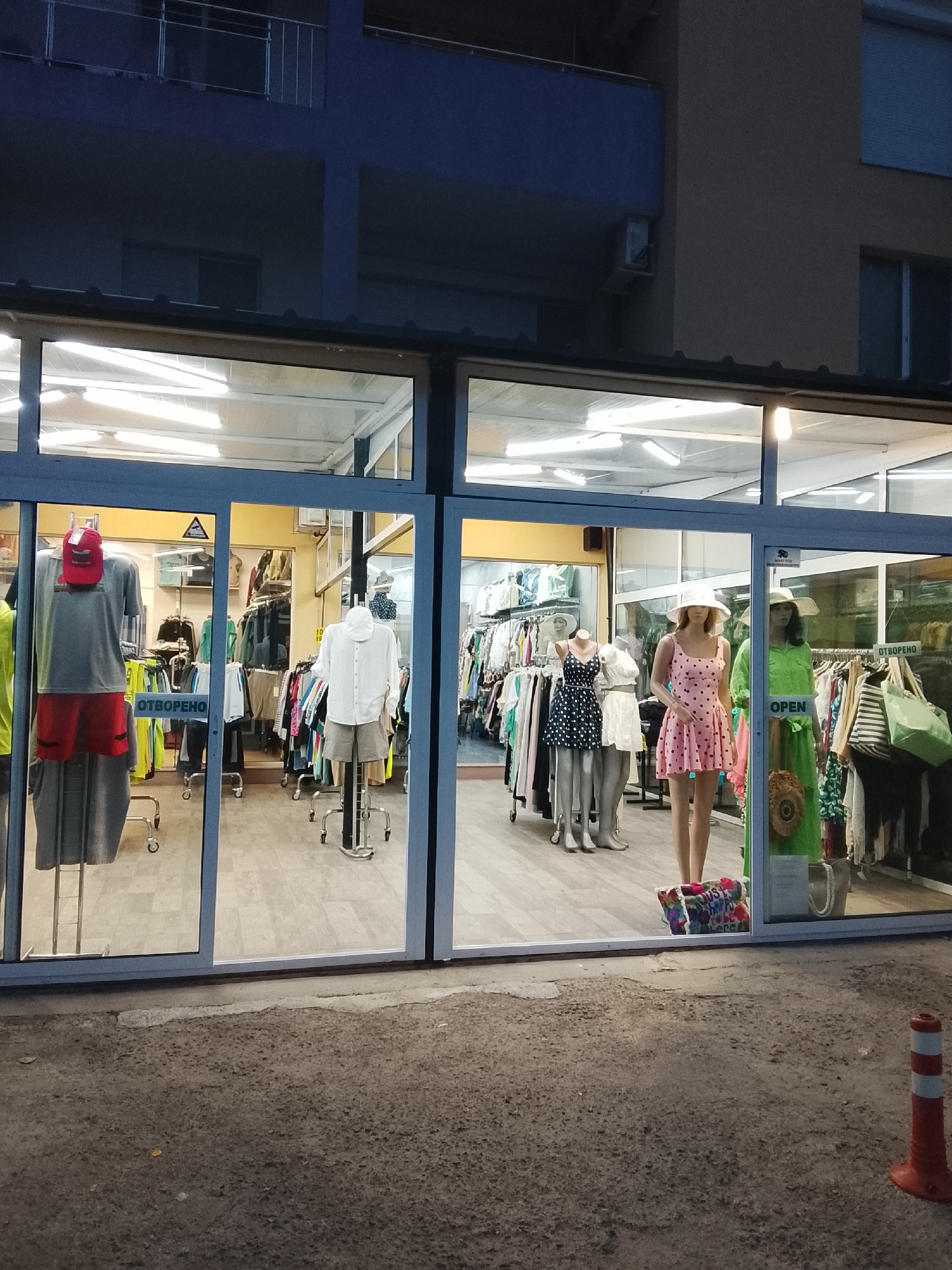 The image shows a storefront in Nesebar, Bulgaria, with mannequins displaying clothing. On the left, a mannequin wears a grey t-shirt, red shorts, and a red cap. Another mannequin is visible behind it. Further inside, clothing racks are filled with various garments. To the right, several mannequins are dressed in summer attire. One mannequin wears a pink polka-dot dress and a white hat, while another to its right is in a green dress with a straw hat. A sign reading "OPEN" is visible on the glass. The store appears to be a clothing boutique. The building is multi-story, with dark blue and beige facade details visible above the storefront. The street in front of the store is paved with asphalt and has a single orange and white traffic cone.