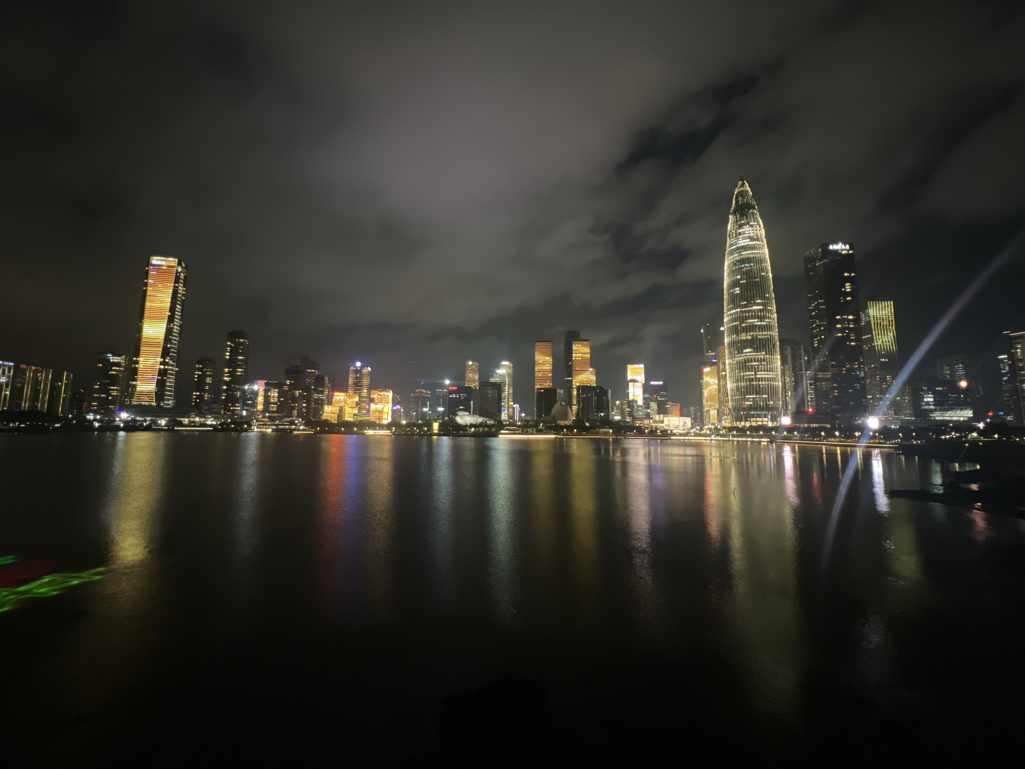 The image depicts a nighttime cityscape of Nanshan District, China, characterized by a dramatic skyline of illuminated skyscrapers reflected in a body of water. The buildings are a mix of modern architectural designs, with some featuring vertical lighting patterns in orange and yellow hues. The most prominent skyscraper on the right side of the image is a tall, slender structure with a pointed top, extensively lit with white lights. The sky above is dark and partially obscured by scattered clouds, which are themselves faintly illuminated by the city lights. The water in the foreground shows a clear reflection of the city lights, creating streaks of color across its surface. On the left side of the water, there are visible green and red laser lights projected onto the surface. No people or animals are discernible in the scene. The overall impression is one of a vibrant, technologically advanced urban environment at night.