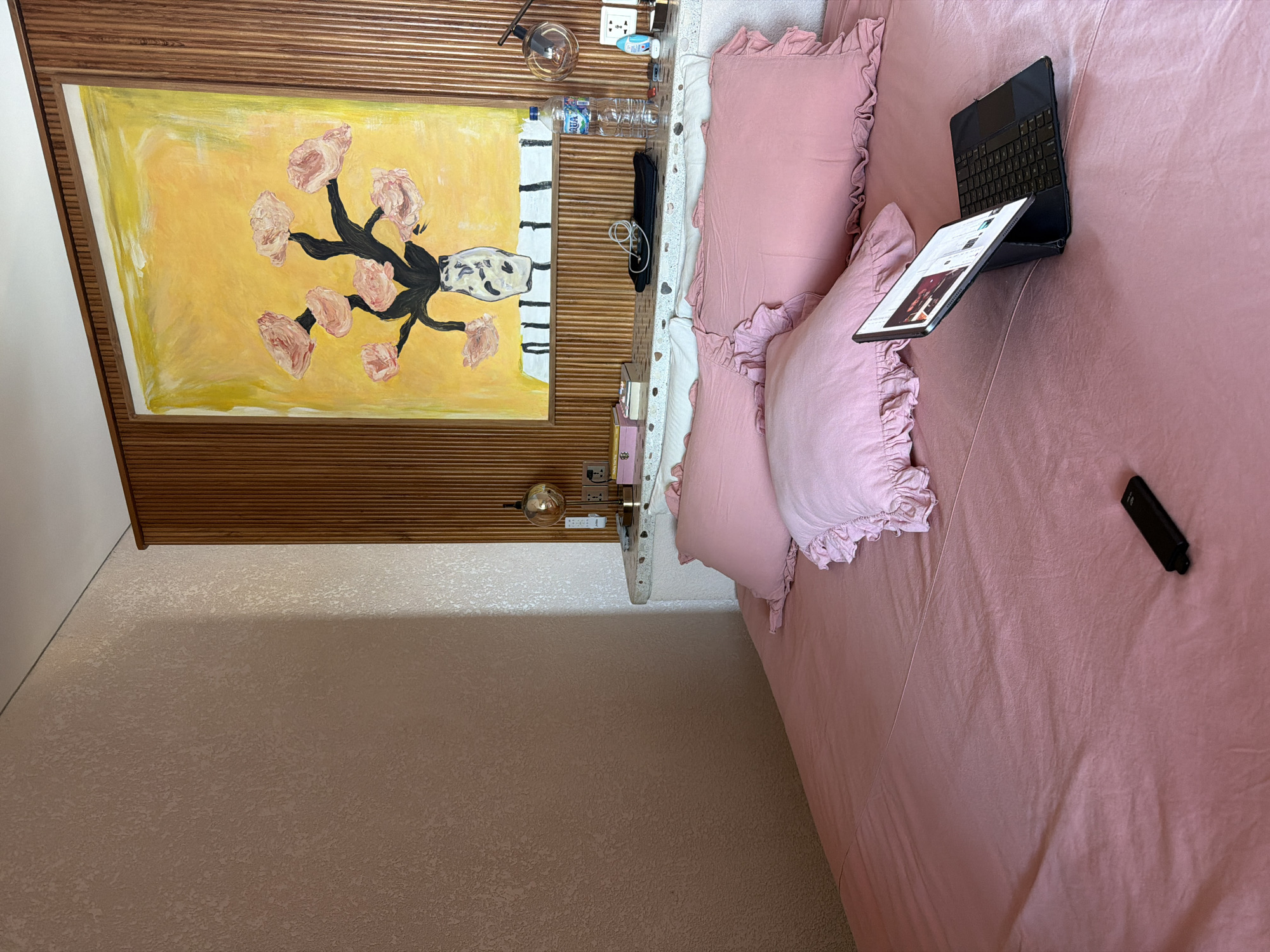 A bedroom scene is depicted, viewed from above, with the image rotated 90 degrees clockwise. The focal point is a bed with pink bedding, including ruffled pillows and a plain pink duvet or sheet. On the bed, near the pillows, rests a black tablet or laptop with an attached keyboard case, open and displaying what appears to be a webpage or app with images and text. A black remote control is also visible on the bed further down.
Behind the bed is a wooden slatted headboard or wall panel, extending upwards. A large rectangular painting hangs centrally on this panel, featuring a vibrant yellow background with a vase holding several pink, petal-like flowers on dark stems. To the right of the bed, a white speckled bedside table holds various items: a clear glass lamp with an Edison bulb, electrical outlets, a blue plastic bottle of "Aqua" branded water, a black pencil case or pouch, a white charging cable, small pink boxes, and a small white decorative object or device.
The wall to the left of the bed and the visible floor area both have a light, textured finish, possibly wallpaper or paint. The room is well-lit, suggesting daytime. The setup indicates a functional and comfortable bedroom in Mengwi, Indonesia.