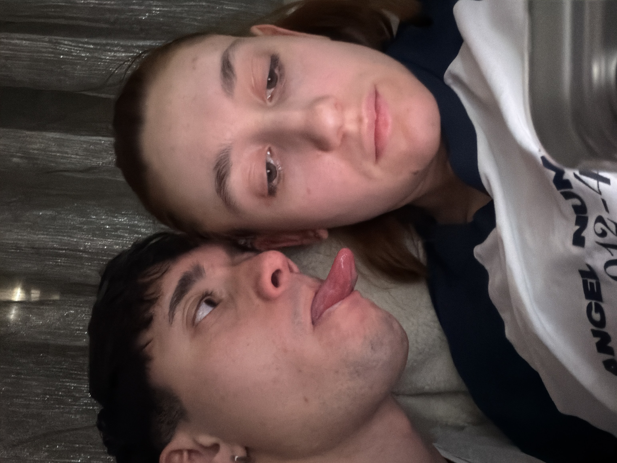 A selfie shows two people lying close to each other. The person on the bottom is a young man with dark hair, wearing a small earring. He is sticking out his tongue and looking upwards. Above him is a woman with light skin and brown hair. She is looking directly at the camera. They are lying on a light-colored surface. The woman is wearing a dark shirt and a white t-shirt with text on it, reading "ANGEL NUMBER 0124". The background consists of horizontal, metallic-looking lines, possibly metal siding or a textured surface. The location and city cannot be determined from the provided image.