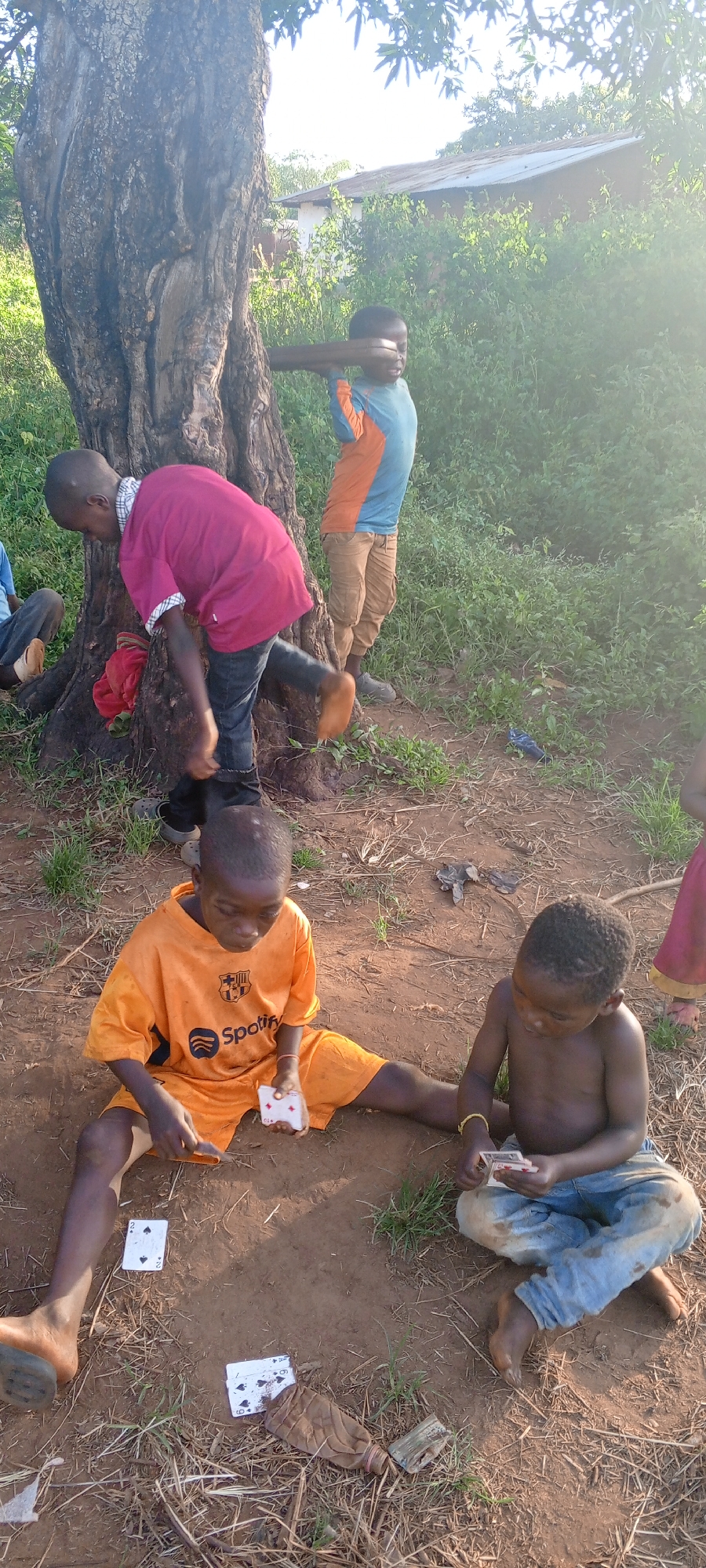In Ngalula, Democratic Republic of the Congo, a group of children are outdoors. One child is leaning against a large tree, another is mid-movement near the same tree, and two shirtless children are seated on the ground, playing with playing cards. Discarded plastic bottles litter the ground. Vegetation and a structure are visible in the background.