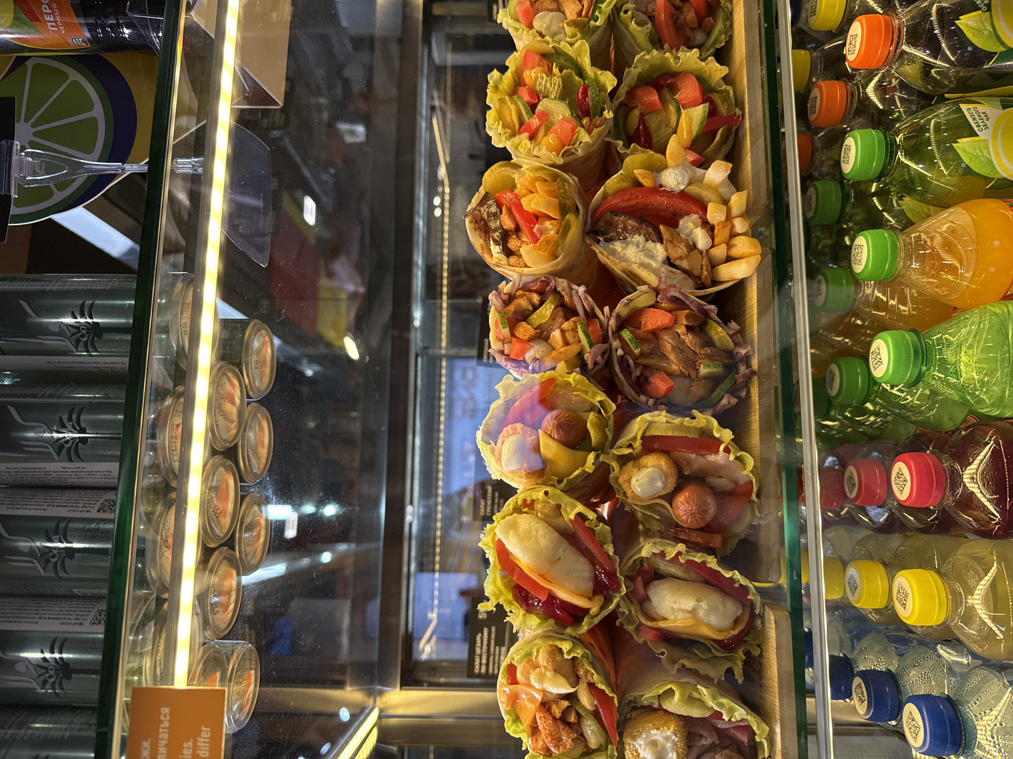 The image depicts the interior of a refrigerated display case in a retail food establishment, specifically located in Khimki, Russia. The scene is dominated by food and beverage items presented for sale.
The upper section of the display case features a horizontal row of multiple identical silver-colored cans, each bearing Cyrillic text and a palm tree-like graphic. Below these cans, an illuminated fluorescent strip light extends horizontally across the frame. An orange rectangular sign, partially visible on the upper left, displays English text "ies. differ" and partial Cyrillic above. To the upper right, a promotional cutout of a lime slice is visible next to a dark-colored beverage bottle.
The central and most prominent section of the display showcases numerous open-faced wraps, arranged in two tiers on a wooden platform. These wraps are formed with outer layers of light green lettuce or cabbage leaves, with their diverse fillings clearly visible. Fillings include various meats (such as chicken, sausages), vegetables (red bell peppers, pickled cucumbers, red onion), yellow cheese, white sauces, red sauces, french fries, and fried potato hash-brown-like pieces. Specific examples include wraps containing chicken pieces, red peppers, and fried potato, and others with small sausages, peppers, and sauces. The items are tightly arrayed. Reflections in the glass behind the wraps show indistinct interior elements and Cyrillic text signs, including "САЛАТ ЦЕЗАРЬ" (Caesar Salad) and "САЛАТ ГРЕЧЕСКИЙ" (Greek Salad).
The lower section of the display contains multiple clear plastic beverage bottles arranged vertically. These bottles have screw caps of various colors, including blue, yellow, red, orange, and green. Each cap displays a small square QR code or similar label. The bottles contain liquids of different colors, such as clear, yellow, orange, red, and green. Condensation is visible on the exterior of several bottles, indicating the refrigerated environment. Some bottle labels are partially visible with Cyrillic text, such as "СВЕЖЕСТЬ ЗЕЛЕНОГО ЧАЯ" (Freshness of Green Tea). No people are visible within the frame.