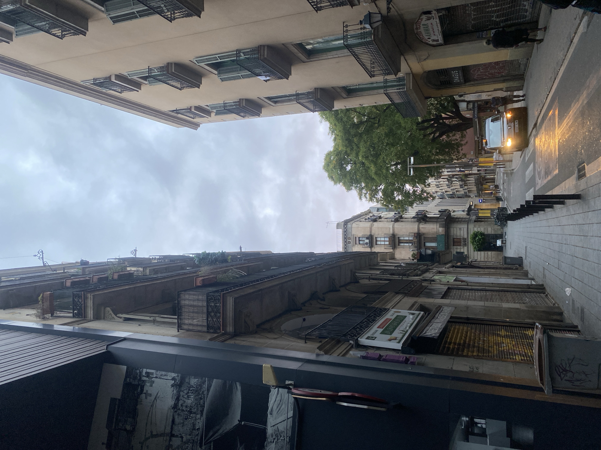 This image captures a street scene on Carrer de la Reina Amàlia in the El Raval neighborhood of Barcelona, Spain, under an overcast sky. The view is angled slightly upwards, looking down a narrow urban street flanked by multi-story buildings, typical of older city architecture.
On the left side of the street, a dark grey refuse bin with graffiti rests on the sidewalk in the foreground. Adjacent to it, a modern dark-paneled building column partially obscures a sign for "PRESTON'S PIZZAS & BURGERS." Further down, a light-colored building with multiple balconies features closed metal roll-down shutters, some defaced with graffiti. A prominent green sign with Arabic script and "ANWAR" (identified as "Carnicería Halal Anwar") is affixed to this building above a shutter. Upper floors display wrought-iron balconies, some adorned with potted plants. In the distance, more residential buildings line the curving street.
On the right side, a tall, light-tan building dominates the frame, extending from the mid-ground to the top right. It features numerous balconies with black wrought-iron railings and dark green wooden shutters, many of which are open. A vintage-style street lamp is mounted on its facade. Below, a closed metal roll-down shutter is marked with "ONAGUS" in white graffiti.
The asphalt street appears wet in places, reflecting the bright yellow light from the headlights of a dark-colored van (possibly a Citroën Berlingo) parked towards the right side of the road in the mid-ground. The van's headlights are illuminated. Two individuals stand near the van: one, wearing a dark top and light trousers, is on the driver's side; the other, in dark clothing, is closer to the passenger side. A third individual, dressed in dark clothing, walks away from the camera along the right-hand sidewalk. The sidewalks are paved with light-colored square tiles, and a line of black bollards separates the left sidewalk from the road. A large green tree is visible above the buildings and van in the mid-distance. A single piece of white litter is visible on the left sidewalk near the foreground.