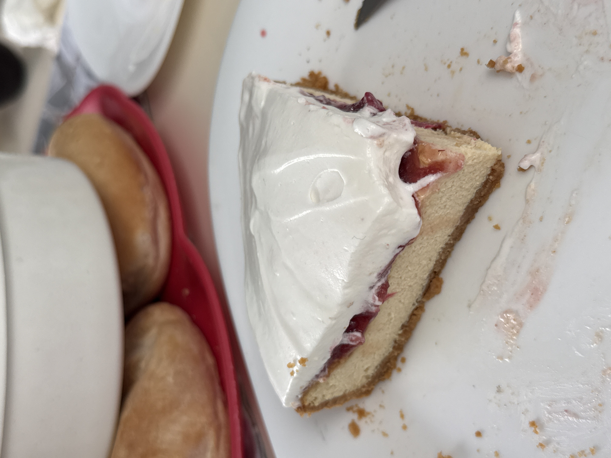 A partially eaten slice of cheesecake sits on a white plate. The cheesecake has a graham cracker crust, a creamy filling, and a thick layer of white topping, possibly whipped cream. There is evidence of fruit filling, possibly cherry, visible between the filling and the topping. Crumbs are scattered around the plate. Next to the plate, a red container holds what appears to be two bread rolls. Parts of a white dish are also visible next to the red container. The scene is an indoor food setting. No people are visible. There is no location information beyond the image itself. The location information provided is not discernible from the image.