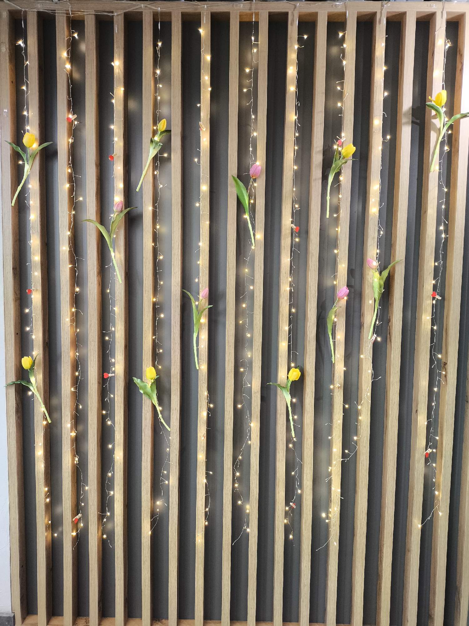 A vertical wall decoration features alternating light-colored wooden slats and dark gray vertical panels. Strings of small, warm-toned fairy lights are draped along the wall, adding illumination. Interspersed among the lights are yellow and pink tulips, appearing to be attached to the wall, adding a touch of color and nature to the scene. Small red objects, potentially decorative elements, are also present along the light strings. The location is not indicated in the image.