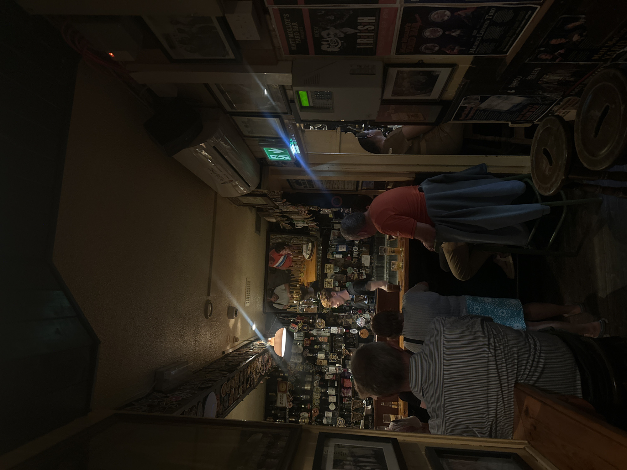 The image depicts the interior of Matt Molloy's Yard Bar in Westport, Ireland, a traditional Irish pub. The dimly lit atmosphere is characteristic of such establishments, with warm lighting from a pendant lamp above the bar casting shadows. The bar itself is well-stocked with numerous bottles of alcohol, neatly arranged on shelves behind the bar. Several patrons are visible, some standing at the bar and others seated. A bartender, a young man with reddish-blond hair, is serving customers. The walls are adorned with a variety of framed pictures and posters, contributing to the pub's character. An air conditioning unit is mounted high on the wall, and an illuminated green emergency exit sign is visible. The overall scene conveys a lively yet intimate pub environment.
