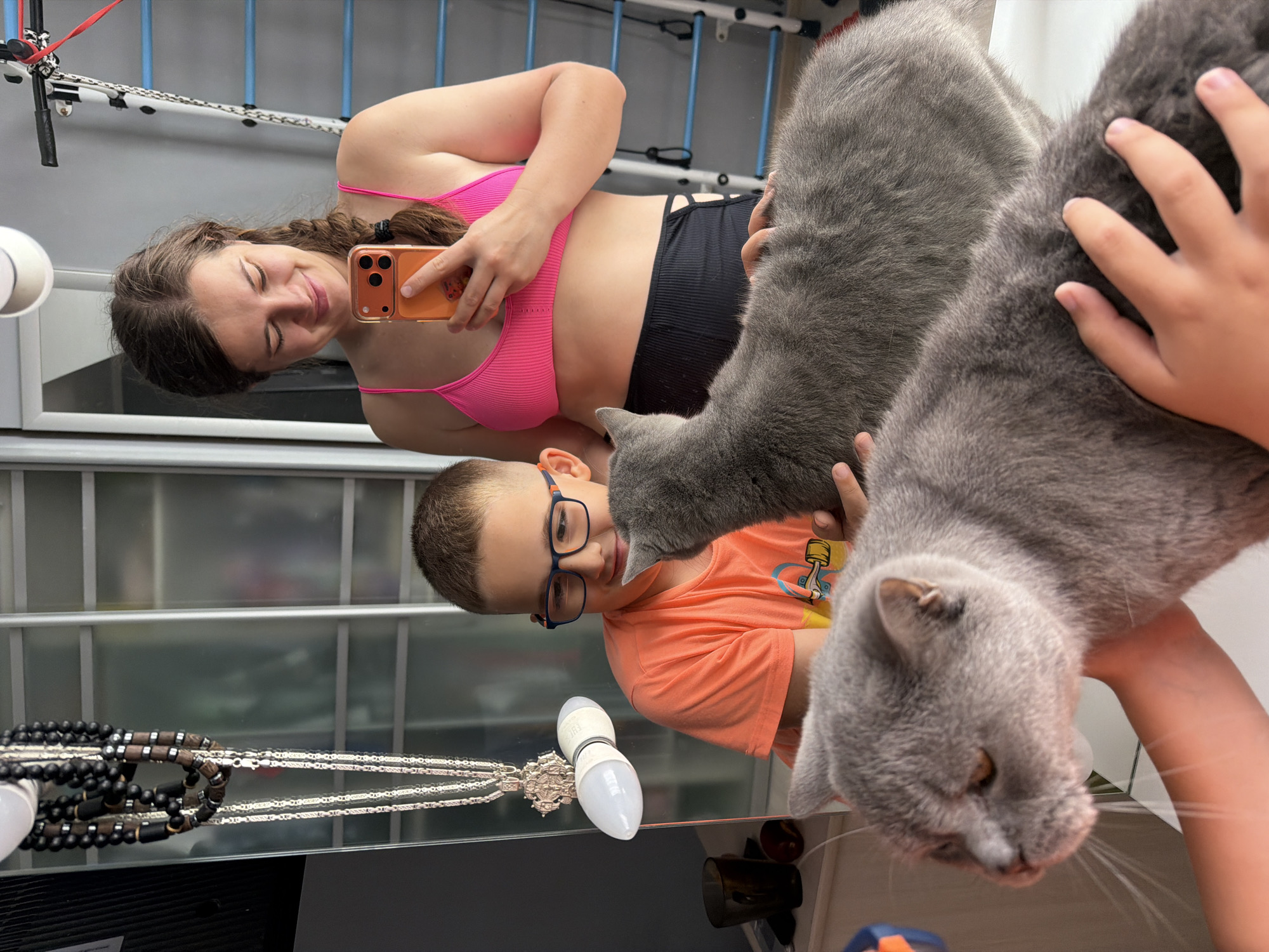 This mirror selfie captures a playful domestic scene involving a young woman, a young boy, and two grey cats, likely taken in Sochi, Russia. The image is rotated 90 degrees counter-clockwise.
In the upper left, a young woman with brown braided hair is seen smiling, holding an orange-cased smartphone to take the photo. She wears a bright pink ribbed bikini top and black high-waisted shorts, reflecting a casual indoor setting.
In the lower-left to central part of the image, a young boy with short brown hair and blue-rimmed glasses is visible, also smiling. He wears an orange t-shirt with an indistinct graphic and appears to be interacting with the cats.
Two fluffy grey cats, likely British Shorthairs, dominate the center and right side of the frame. One cat is held by the boy, looking towards him, while the head of another cat is prominently featured in the lower right, possibly being petted or held by another hand (or the boy's other hand).
The background, reflected in the mirror, shows white and grey elements that resemble shelving or a structural frame, indicating an indoor environment. On a surface in the lower left, a necklace with black beads and silver chains/pendants is visible next to a white light bulb.
Both the woman and the boy display cheerful expressions, suggesting a happy and relaxed atmosphere. No clear text is visible on any objects. The overall impression is a candid moment of family and pet interaction.