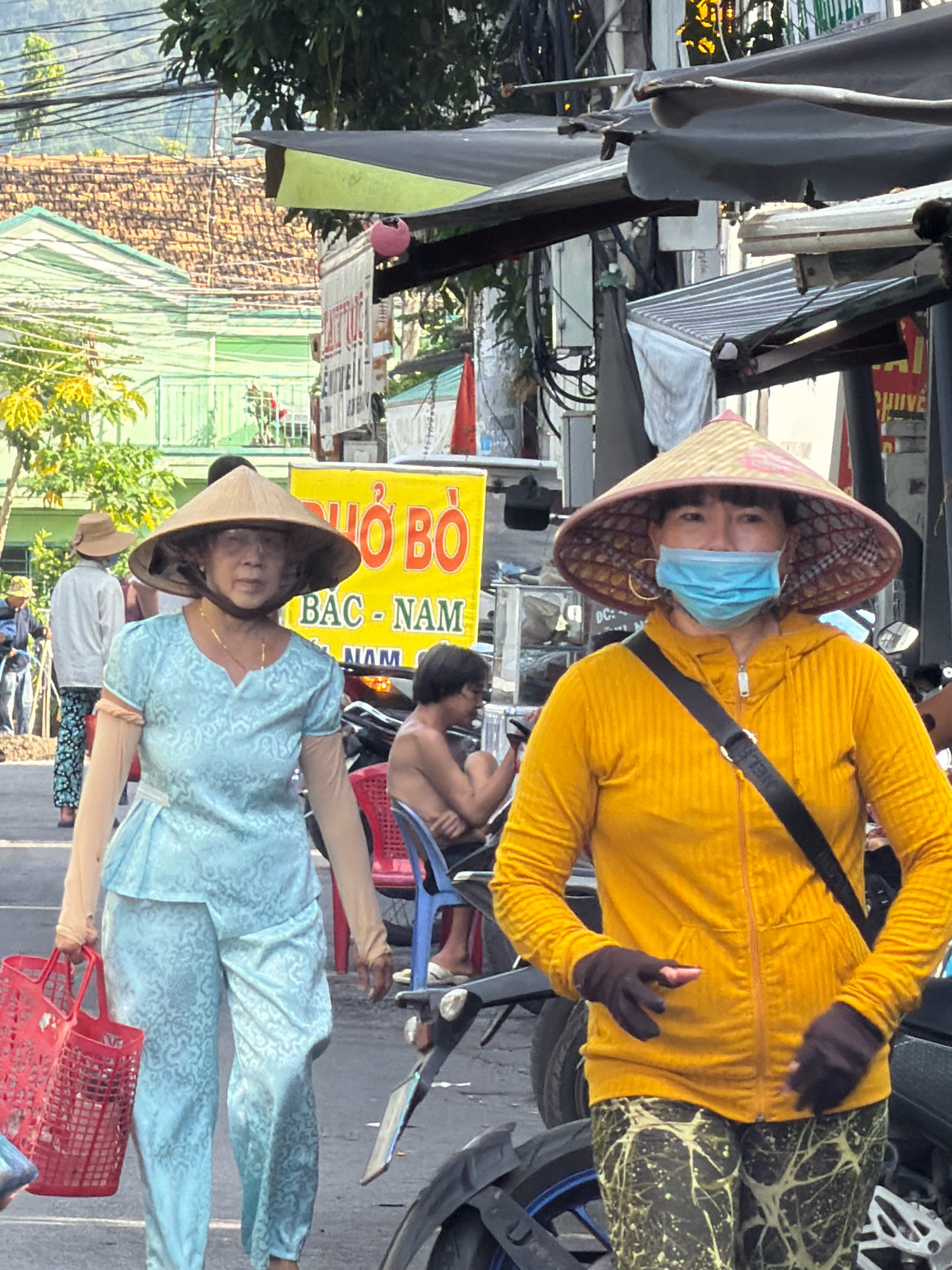 Two Vietnamese women are walking down a street in Nha Trang, Vietnam. The woman on the left is wearing a light blue patterned outfit and a conical hat. She is carrying a red basket. The woman on the right is wearing a yellow hooded jacket, patterned leggings, and a conical hat with a blue face mask. She is also wearing dark gloves. In the background, there are buildings, trees, and other people. The scene appears to be a marketplace or a busy street.