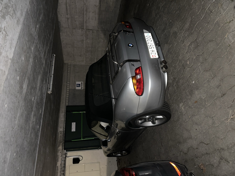 A silver BMW Z3 convertible with a black soft top is parked facing inwards in an underground parking garage in Bassersdorf, Switzerland. The vehicle features a chrome luggage rack on its trunk lid, a "Z3" badge, a BMW emblem, and a Swiss license plate reading "ZH-557200". Below the license plate are twin exhaust pipes and a visible trailer hitch. The car's left rear is positioned next to the right rear of a partially visible dark-colored vehicle. The surrounding environment consists of rough concrete walls and ceiling. A dark green metal door or storage unit is visible behind the BMW. An overhead fluorescent light fixture illuminates the scene. A white sign with a blue "P" and the text "Nur für Gäste" (Only for Guests) is affixed to the wall. The concrete floor shows tire marks. No individuals are present, and no actions or interactions are depicted.