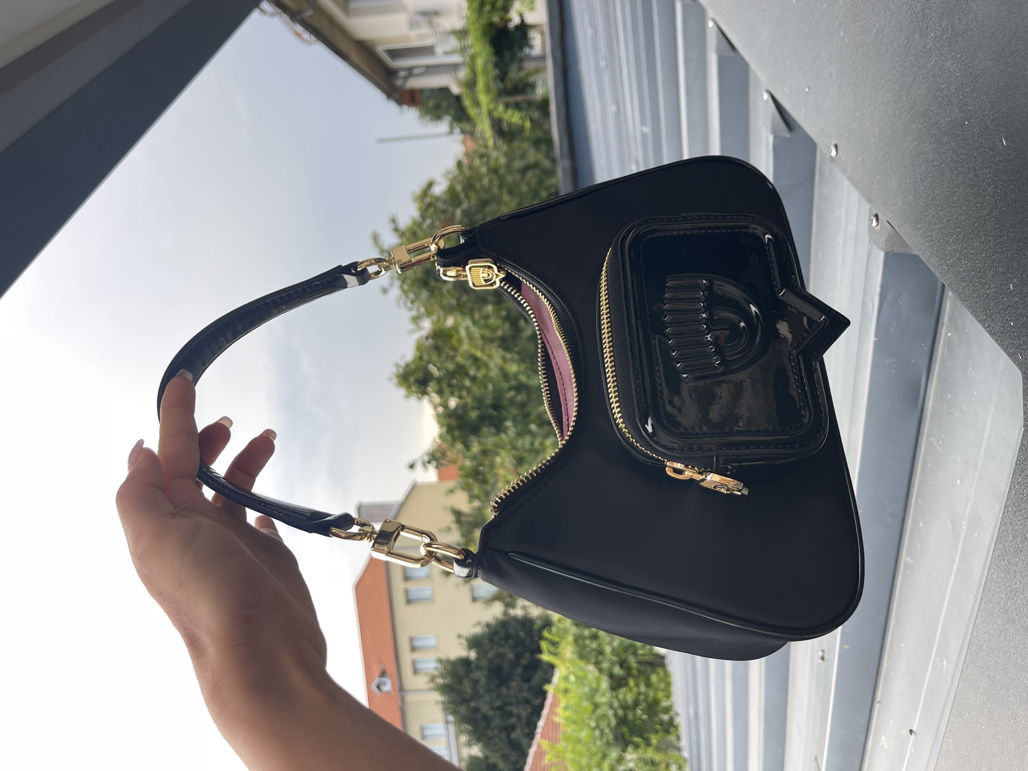 A hand with light-colored skin and manicured nails holds a black shoulder bag. The bag has a patent leather front pocket with a distinctive logo that resembles an eye. Gold-toned hardware connects the strap to the bag. The background consists of a cloudy sky and buildings with orange roofs, suggesting an outdoor urban setting. The scene appears to be captured from a slightly elevated position, possibly a balcony or rooftop, overlooking a street in Karlovo, Bulgaria.