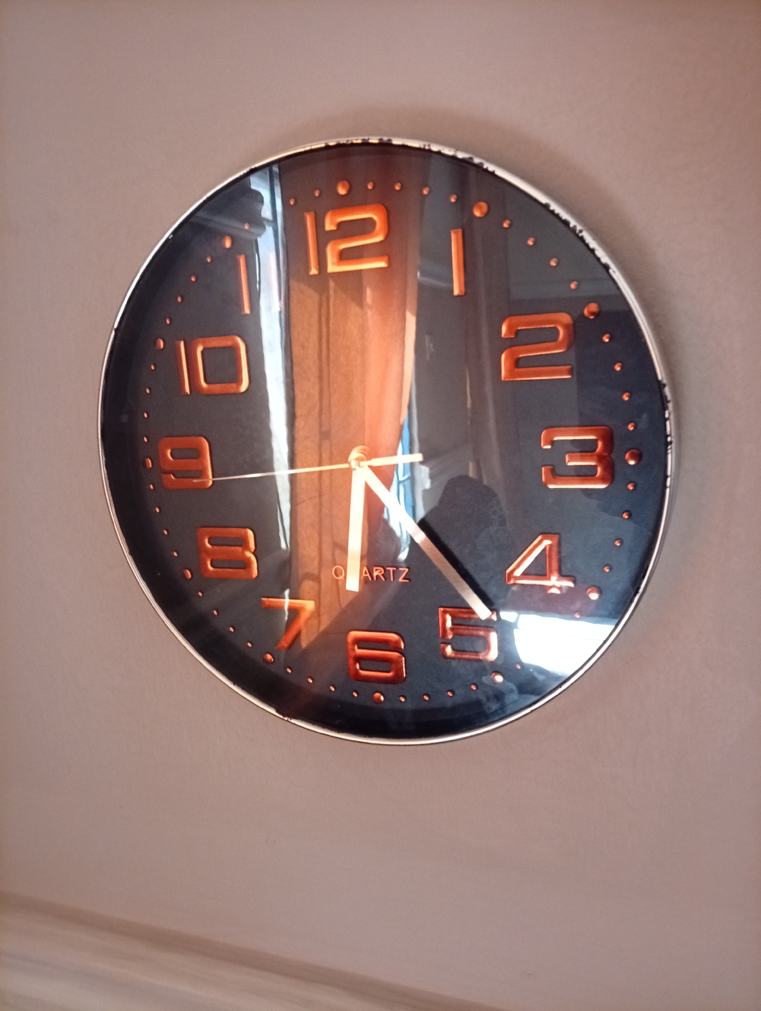 The image shows a close-up of a dark-faced wall clock with orange numerals and hands. The clock is positioned on a neutral-colored wall. The numerals are large and modern in design, with the numbers 12, 2, 3, 4, 5, 6, 7, 8, 9, and 10 clearly visible. The clock hands appear to be white. A reflection on the clock face suggests a window or light source is present, and a blurred figure, possibly a person, can be discerned in the reflection. The clock face also has small orange dots marking the hours. The clock has the word "QUARTZ" printed on it. There are no people or identifiable objects in the environment other than the clock itself. The image does not contain any information that could link it to Gura Top, Nigeria or any specific city.