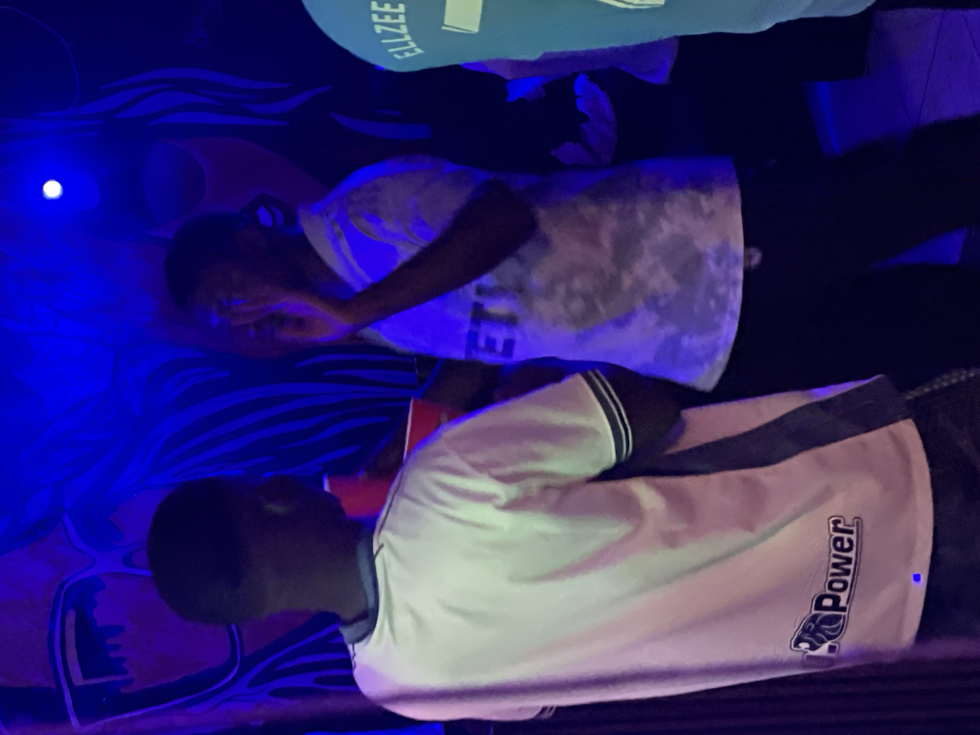 The image depicts an indoor scene with several individuals, predominantly young Black males, socializing in what appears to be a nightclub or party setting. The environment is characterized by prominent blue lighting and a mural or painted backdrop featuring stylized facial features and abstract designs.
In the foreground, a young man wearing a white t-shirt with a dark stripe on the sleeve and a logo reading "Power" is partially visible from the back. To his right, another young man in a light-colored, possibly tie-dye shirt, is facing towards the right and gesturing with his hand near his face. His shirt has text on it that is partially obscured. To the far right, a portion of a third individual's light blue t-shirt is visible, displaying the text "ELLZEE".
The people appear to be engaged in conversation or enjoying the ambiance of the location. Red cups are visible, suggesting the presence of beverages. The overall impression is one of a lively social gathering. The location within the city of Durumi, Nigeria is not explicitly indicated by any visual cues in the image itself.