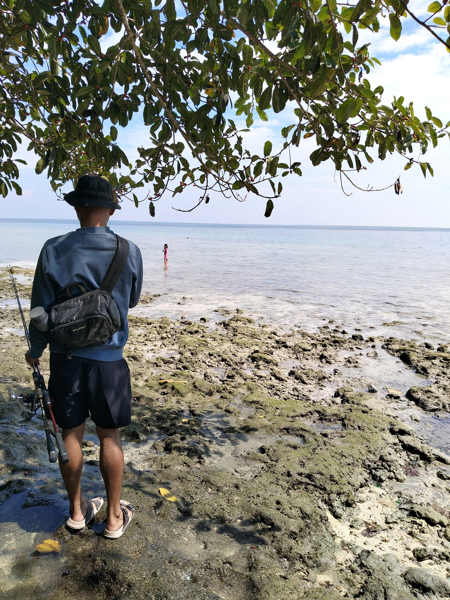 A man wearing a black bucket hat, a blue long-sleeved shirt, and black shorts stands on a rocky, moss-covered shoreline, holding a fishing rod. He has a black crossbody bag slung over his shoulder. In the distance, a child in a pink outfit stands in the shallow water. The sea stretches out to the horizon under a partly cloudy sky, framed by the branches of a leafy tree. The scene appears to be a coastal area in Biak, Indonesia.
