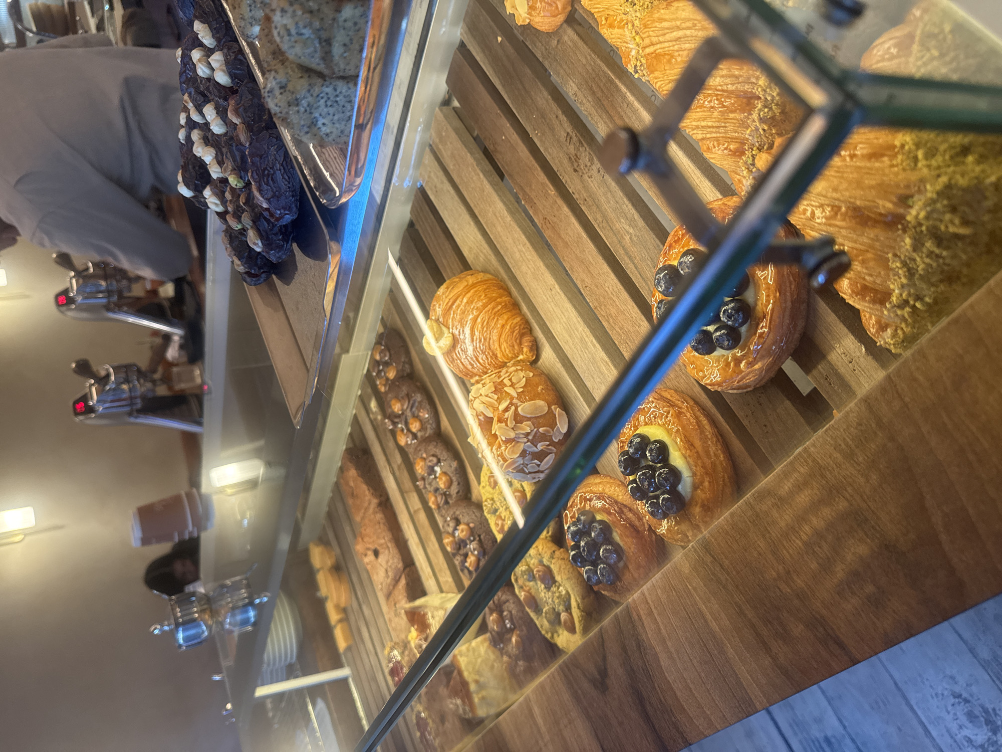 The image shows a display case filled with various pastries, including croissants with almond slices, blueberry danishes, and chocolate pastries. The pastries are arranged on wooden slats inside the glass case. A person wearing a light-colored garment is visible in the background, possibly behind the display. The scene is possibly located in a bakery or cafe in Yerevan, Armenia, although the exact location cannot be determined from the image.