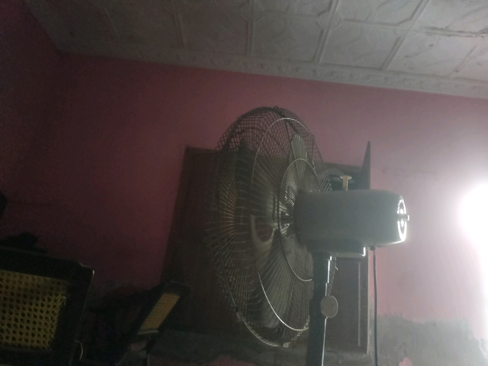 An electric fan is prominently displayed in the foreground, angled towards the left side of the frame. The fan has a metallic grill and its motor housing is visible on the right. To the left of the fan, a portion of a dark object with a yellow, grid-like pattern is visible. The background consists of a pinkish-purple wall with a textured, white ceiling above it. The lighting in the image is uneven, with a bright light source to the far right, casting a strong glare. The scene appears to be indoors. The location is identified as Kot Hammeraj, Pakistan.