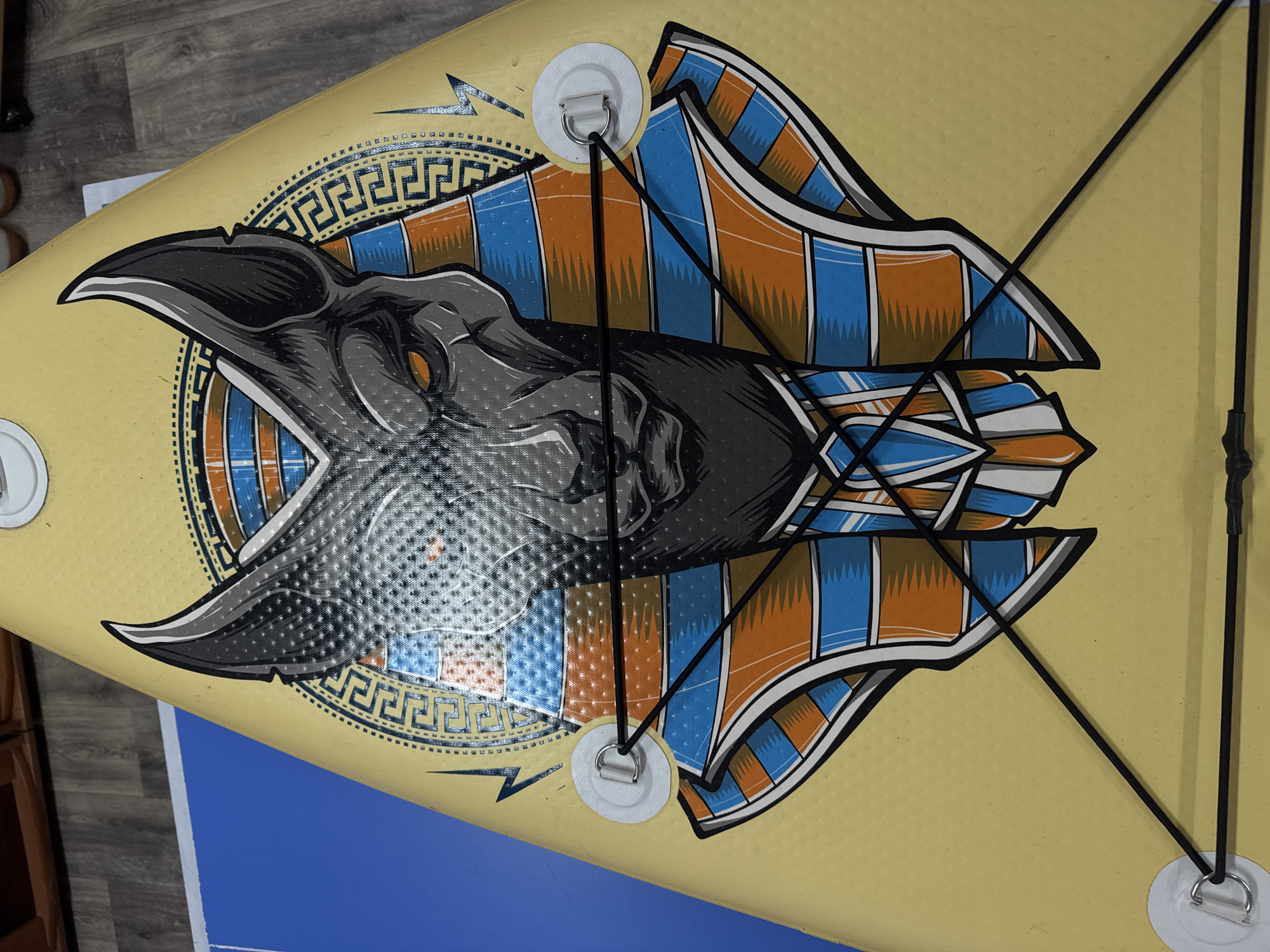 A yellow stand-up paddleboard is seen from above. The paddleboard has a graphic of a bird's head with blue and orange wings. The graphic is surrounded by a Greek key pattern. A black bungee cord is attached to four white D-rings on the paddleboard. The background is a gray wooden floor and a blue surface. The image was taken indoors. The location of Прикамский, Russia, is not evident.