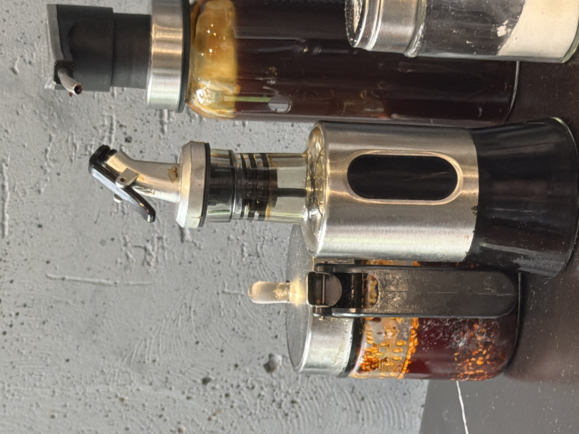 The image shows a close-up of three glass containers, likely for condiments or spices, mounted on a textured gray wall. The top container is filled with a dark brown liquid, possibly oil or vinegar. The middle container is clear with a metallic dispensing nozzle and a stainless steel section with a rectangular cutout. The bottom container is filled with a dark red substance with visible particles, possibly chili oil or pickled spices, with a smaller dispenser. The wall has a rough, textured surface. There is no indication of the location being Saint Petersburg, Russia.