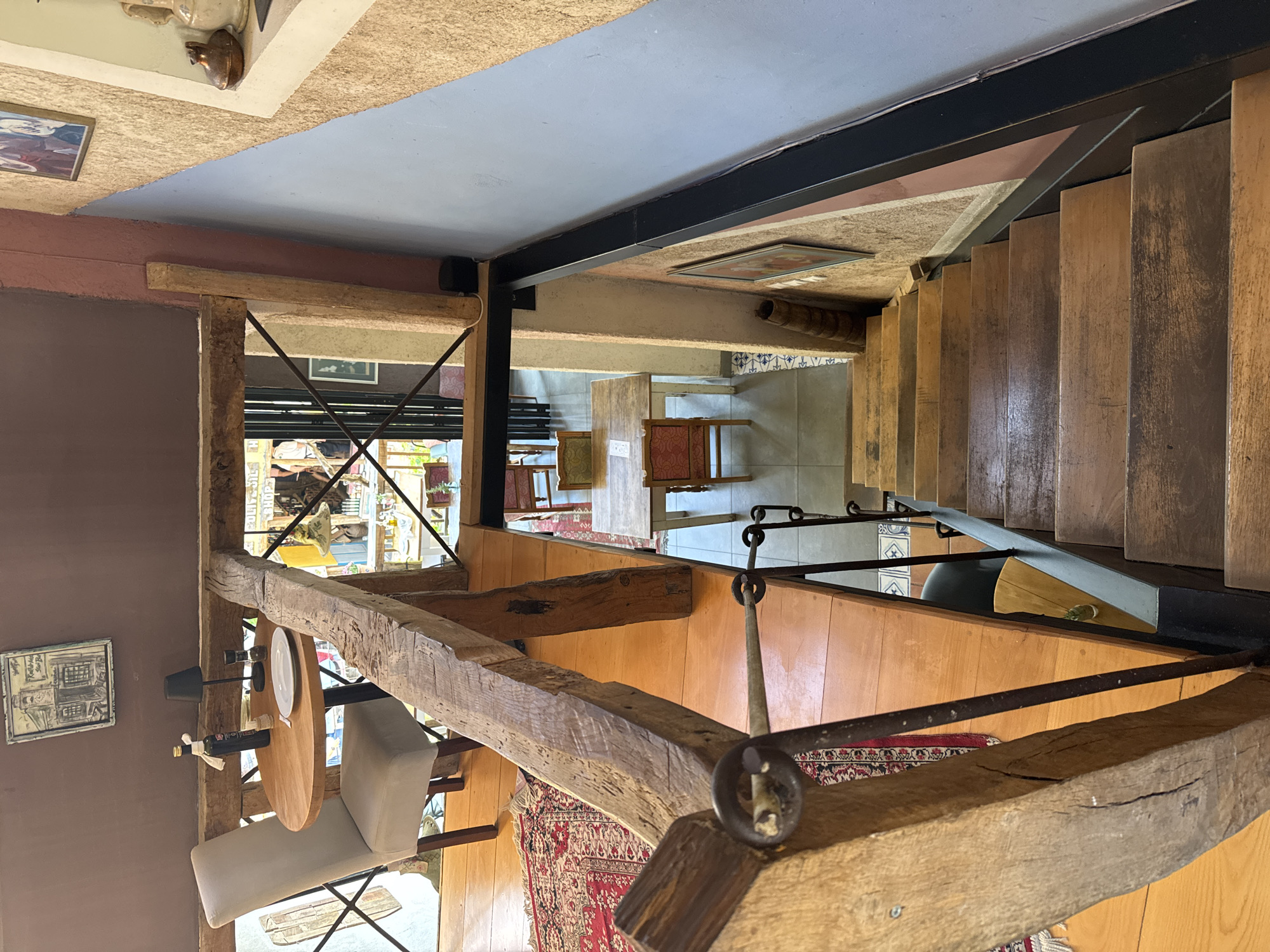 This interior shot depicts an elevated perspective of a room with a blend of rustic and modern elements. A heavy, exposed wooden beam framework is prominent in the foreground, with an attached small round table set for one with a wine bottle and glass. The view extends towards a staircase constructed of wood treads. In the background, a glimpse of an outdoor scene, potentially a street or shop, is visible. A red patterned rug adorns the floor, and the surrounding walls feature a mix of materials, including wood paneling and painted surfaces. The location is likely an interior space within the city of Bartula, Montenegro, identifiable by the distinctive architectural style and decor.