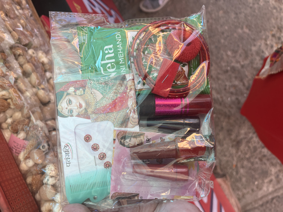 The image shows a plastic bag containing various beauty and decorative items, likely purchased at a market stall in Panchkula, India, located at the coordinates 30.725 latitude, 76.86 longitude. The bag contains two lipstick tubes, a small comb, a pack of bindi, a packet of henna, a small red pom-pom, and two golden bangles. The image is taken from above, with the bag held in someone’s hand. The background is a blurred image of a marketplace.  A woman in a red and green dress and a woman with brown hair and pink clothing are visible in the background.