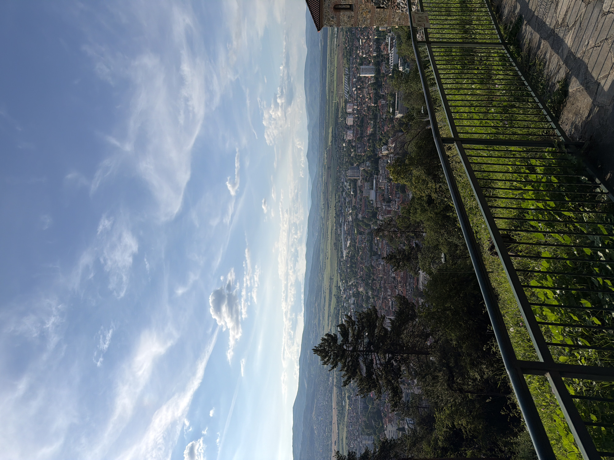 This image presents a high-angle panoramic view of the city of Kyustendil, Bulgaria, captured during daylight under a partly cloudy sky.
In the immediate foreground, a dark green metal railing with vertical balusters extends horizontally across the lower portion of the frame, curving slightly to the right. Behind the railing, lush green grass and various plants, some with broad leaves, cover a slope. A paved stone path, composed of irregular flagstones, is visible on the ground to the right of the railing.
The midground reveals a significant portion of the city of Kyustendil. Numerous buildings, predominantly residential with varied roof colors, are densely packed across the landscape. Several taller, more modern multi-story buildings are interspersed among the older structures. Dark green evergreen trees, notably a large pine tree centrally positioned, rise from the slope, partially obscuring parts of the city. On the far right, a section of a stone building with a reddish-brown tiled roof and a small arched window is visible.
In the background, the urban area transitions into expansive green fields and agricultural land. Beyond these fields, a long chain of undulating hills and mountains stretches horizontally across the horizon, appearing hazy blue in the distance. The sky above is a bright, clear blue, adorned with numerous scattered white clouds, ranging from wispy cirrus formations to more defined cumulus clouds. No people or animals are visible in the scene.